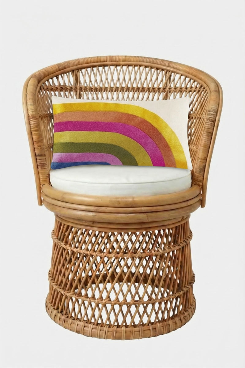 A wicker chair with a round white cushion features the Calistoga Embroidered Rainbow Lumbar Pillow, showcasing yellow, pink, green, and blue embroidery on a white background. The chair is set against a plain white backdrop.