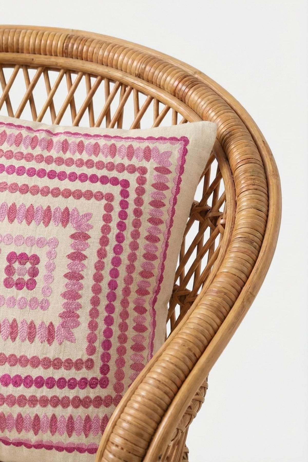 Close-up of a rattan chair with the Carmel Embroidered Pillow 20x20", showcasing a bold pink and red geometric pattern of dots and lines forming concentric squares on a cream background.