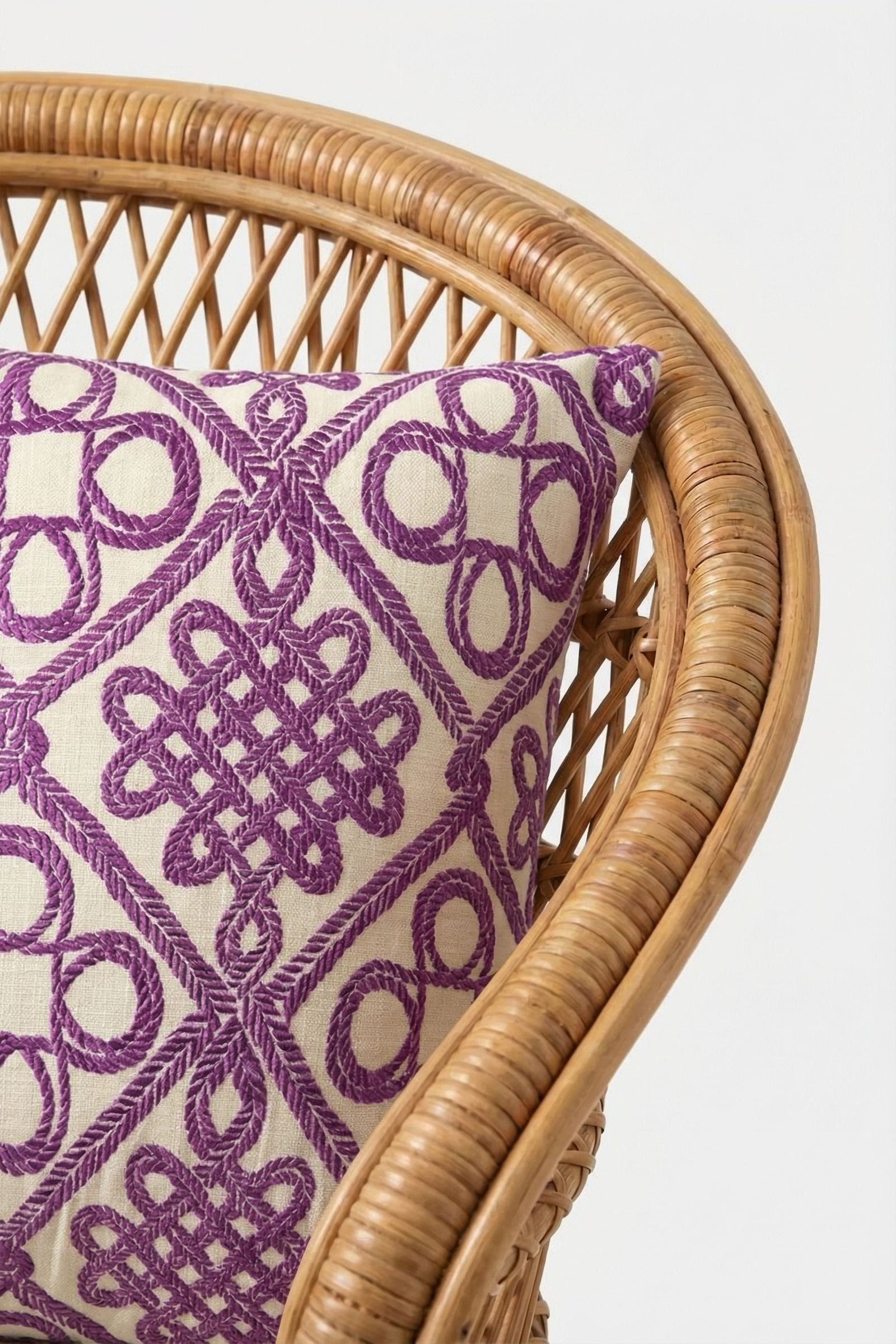 A close-up of a wicker chair with the Costa Mesa Embroidered Throw Pillow, showcasing its intricate purple geometric pattern on a cream background.