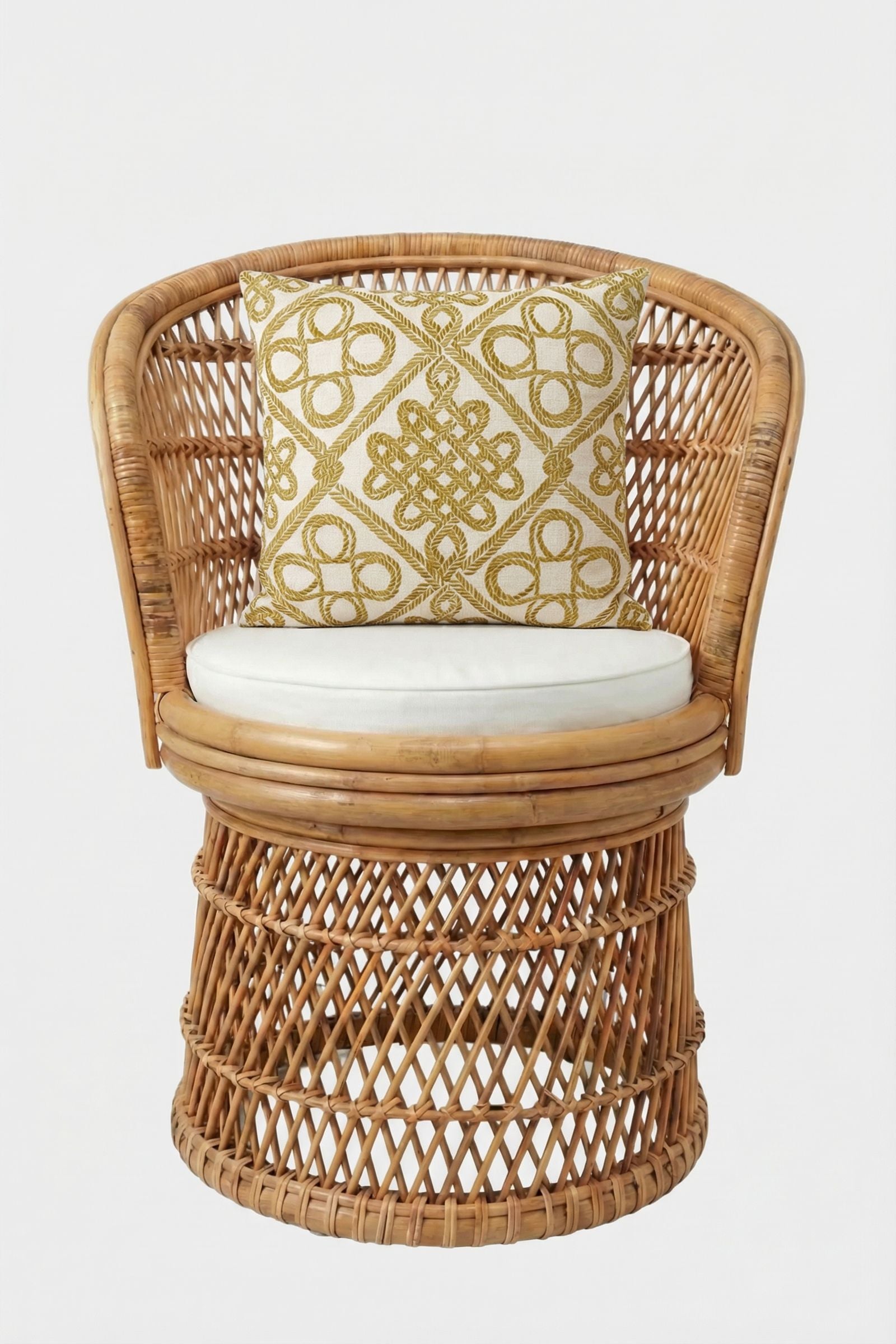 A round wicker chair with a curved back and white seat cushion is accented by the Costa Mesa Embroidered Throw Pillow, featuring a gold geometric pattern on white. The setup is against a plain white backdrop.