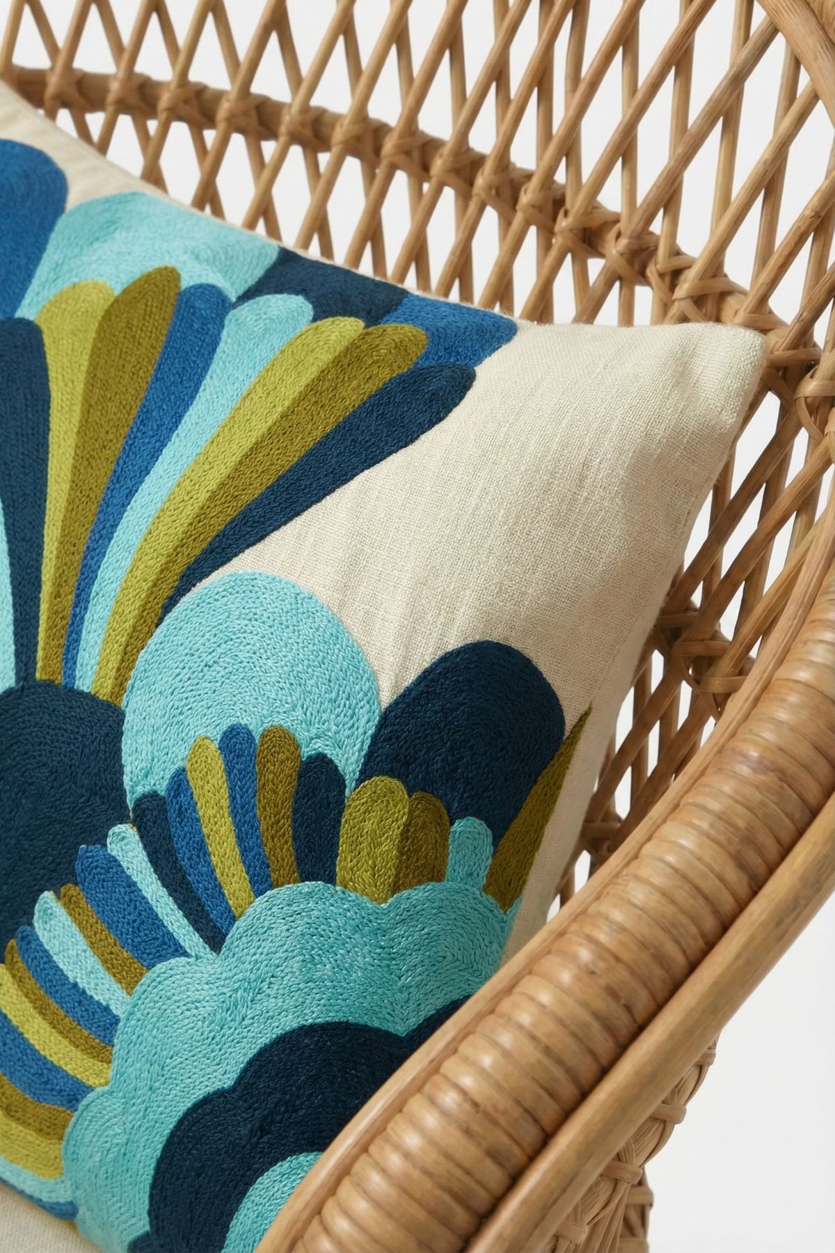 A close-up of the Escondido Embroidered Lumbar Pillow on a rattan chair, showcasing its beige cover with bold blue, teal, green, and navy abstract embroidery. The background is white.