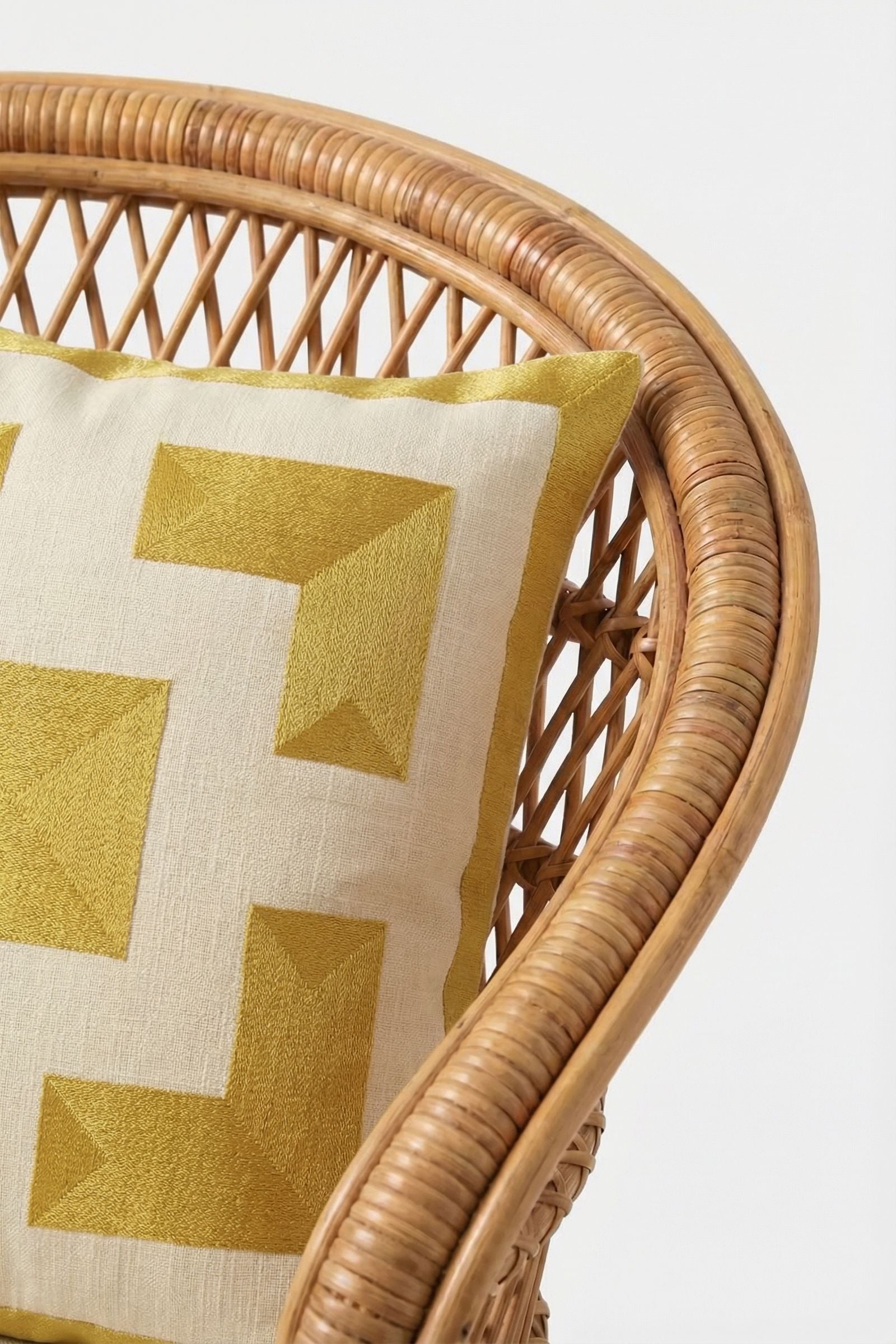 Close-up of a curved rattan chair with the Palm Springs Block Embroidered Pillow, a 20"x20" off-white cover with a bold geometric yellow design and plush down feather cushion, set against a plain white background.