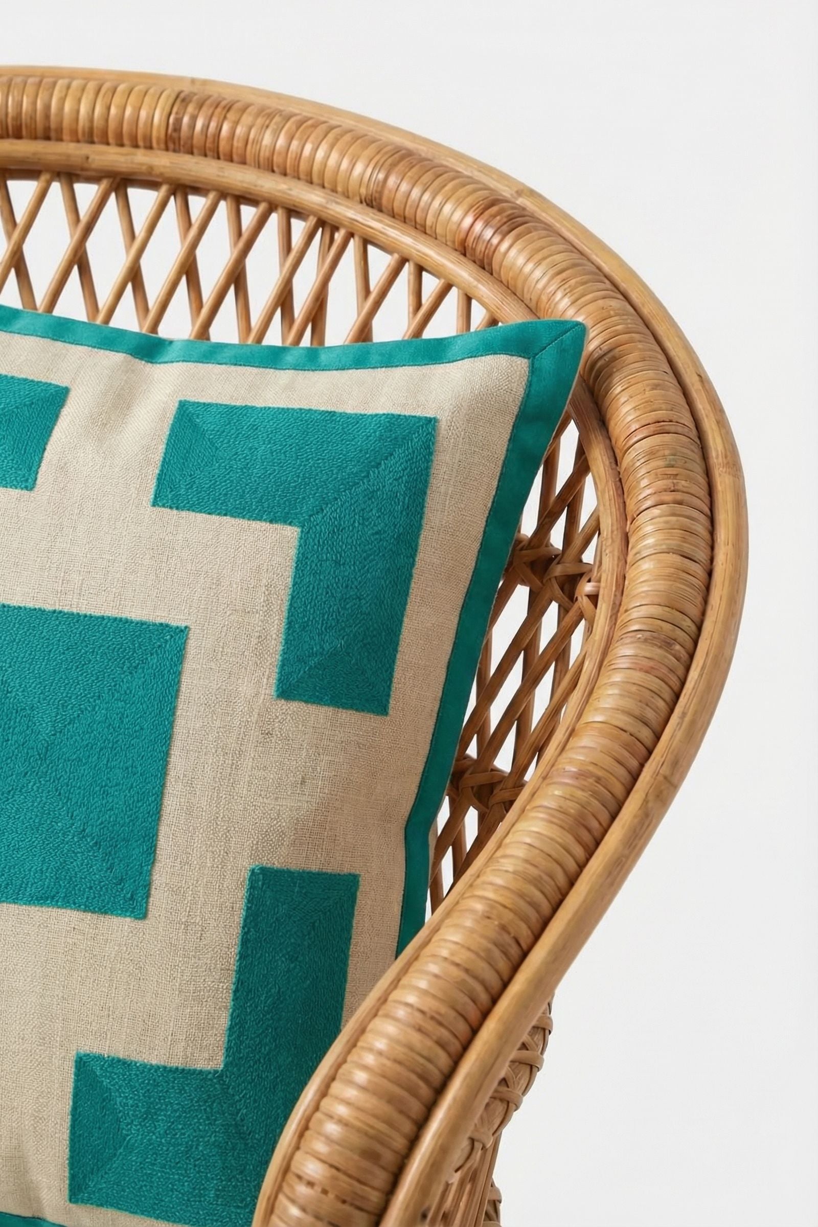 A close-up of a rattan chair featuring the Palm Springs Block Embroidered Pillow (20"x20") in beige and teal, filled with plush down feathers, displayed against a plain white background.