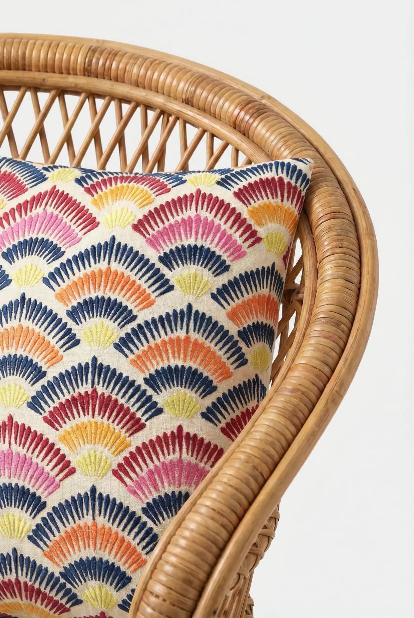 A close-up of a rattan chair with the Pasadena Embroidered Throw Pillow, showcasing a colorful fan or scallop pattern in red, orange, yellow, and navy blue on a white background.