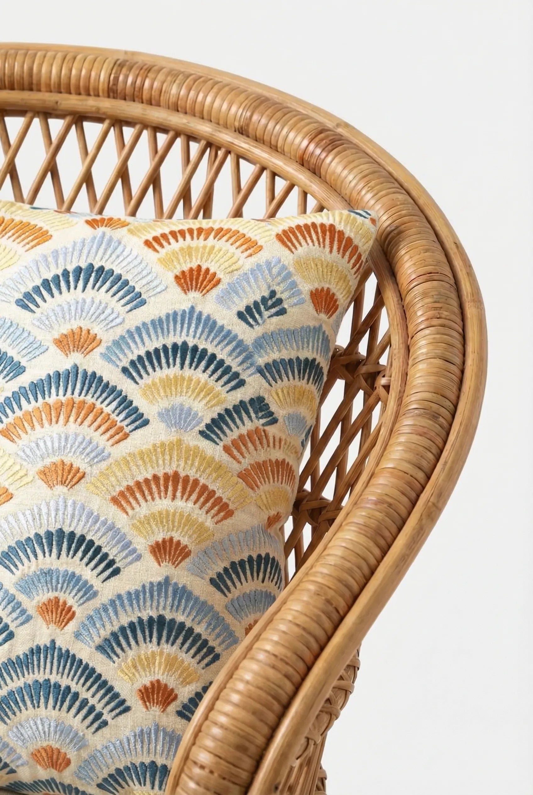 A close-up of a woven rattan chair with the Pasadena Embroidered Throw Pillow, featuring a colorful fan-shaped geometric pattern in blue, yellow, and orange tones.