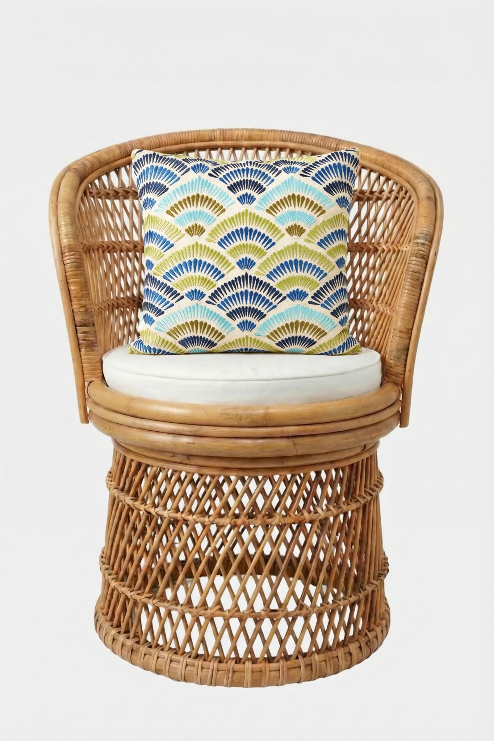 A round wicker chair with a white cushion is accented by the Pasadena Embroidered Throw Pillow, which displays a blue, yellow, and white fan pattern, all set against a plain white background.