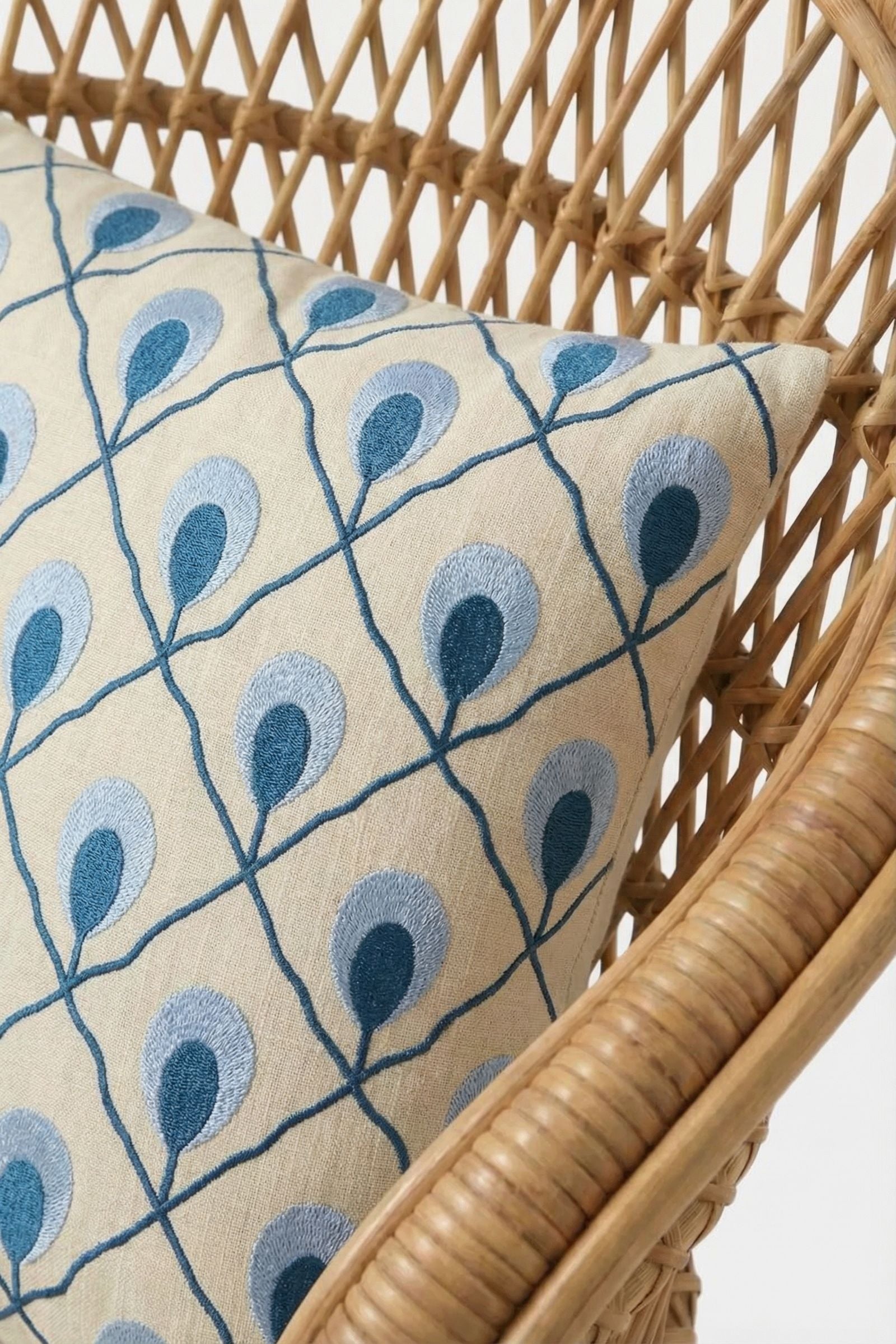 A close-up of a rattan chair with the Roseville Embroidered Lumbar Pillow, featuring a beige cushion with blue and teal embroidery in a peacock feather or leaf pattern, set against a plain white background.