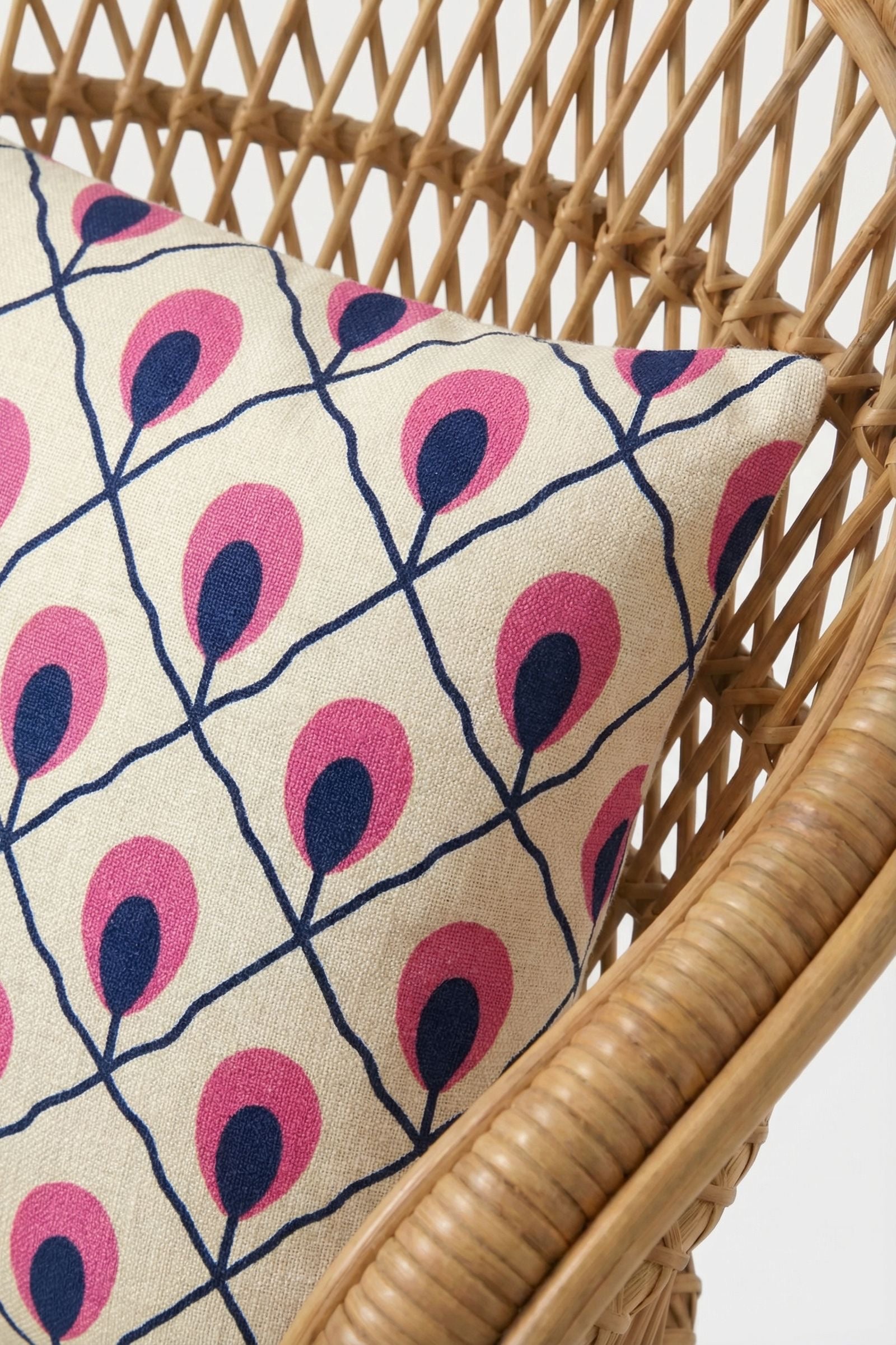 The Roseville Embroidered Lumbar Pillow, featuring navy blue lines and pink leaf-like accents in a geometric pattern, rests on a light brown woven rattan chair.