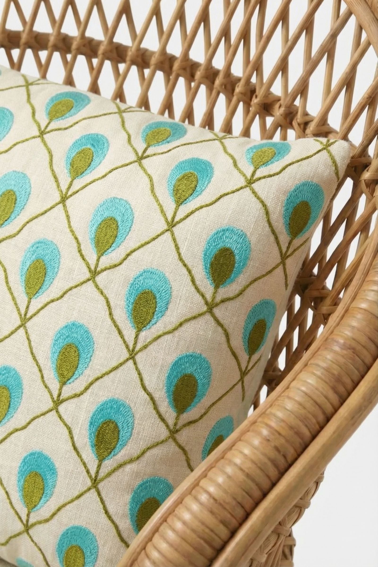 The Roseville Embroidered Lumbar Pillow, cream-colored with blue and green peacock feather designs, is shown on a light brown woven rattan chair.