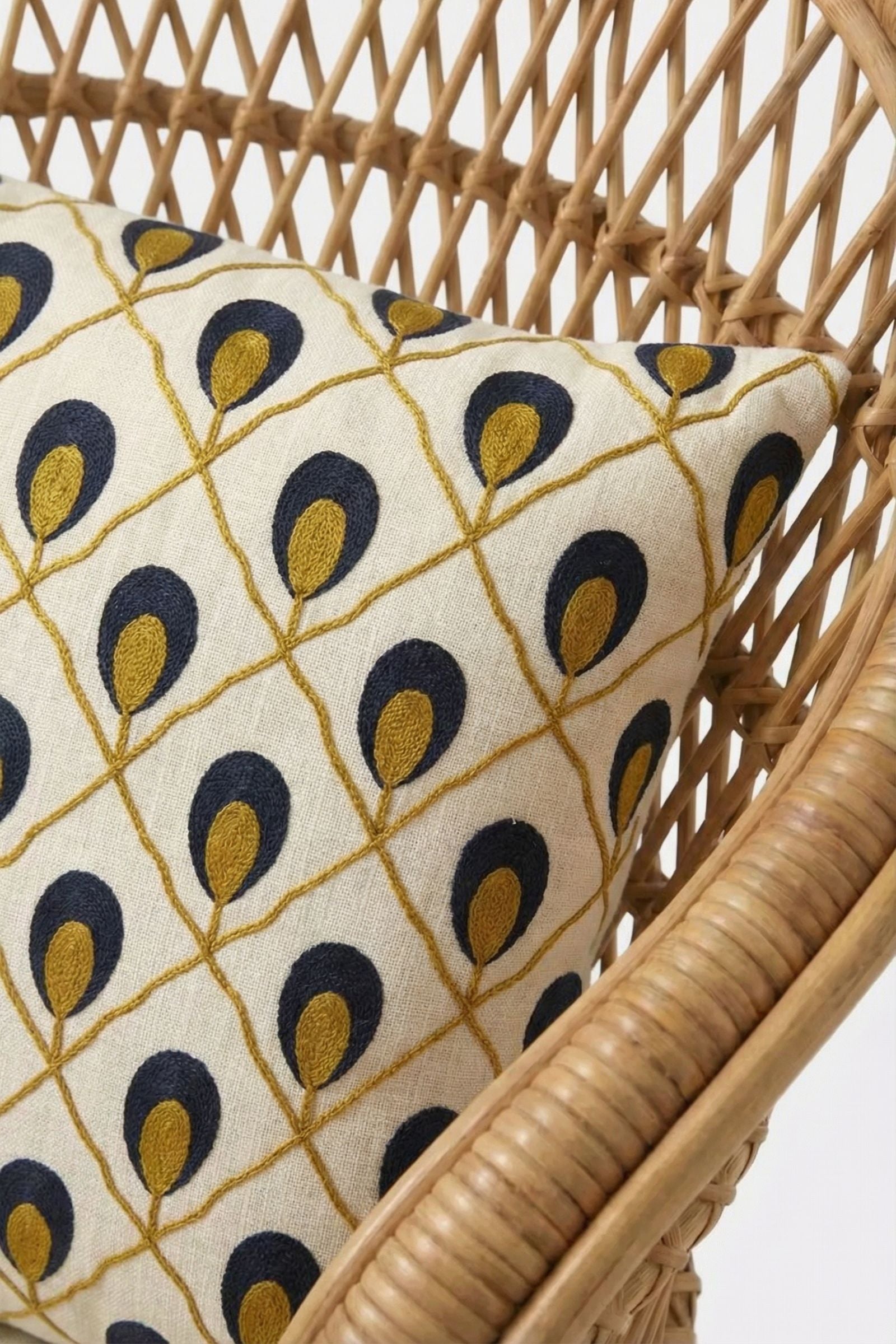 A close-up of a woven rattan chair with the Roseville Embroidered Lumbar Pillow, which features a geometric pattern of navy blue and mustard yellow teardrop shapes on a cream background.