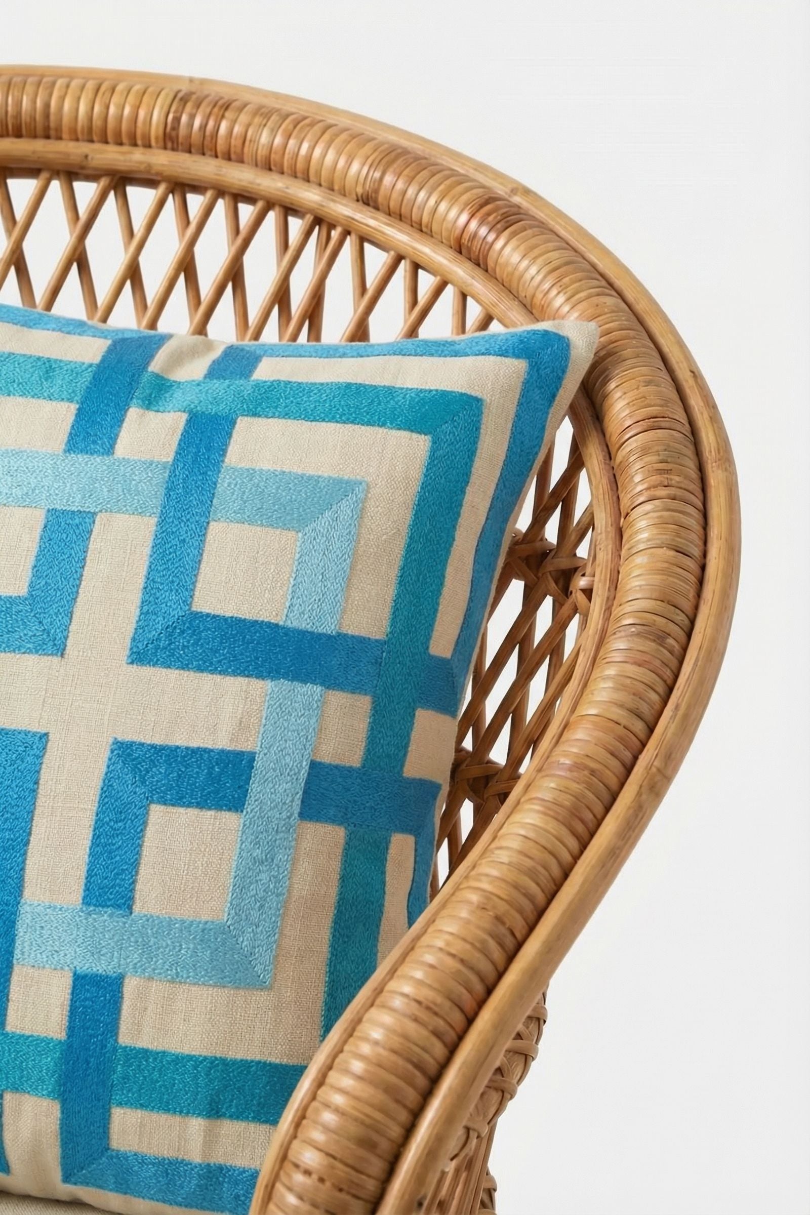 A rattan chair with a beige Shanghai Links Embroidered Throw Pillow, showcasing a modern blue geometric pattern, stands out for its bold design against a plain white background.