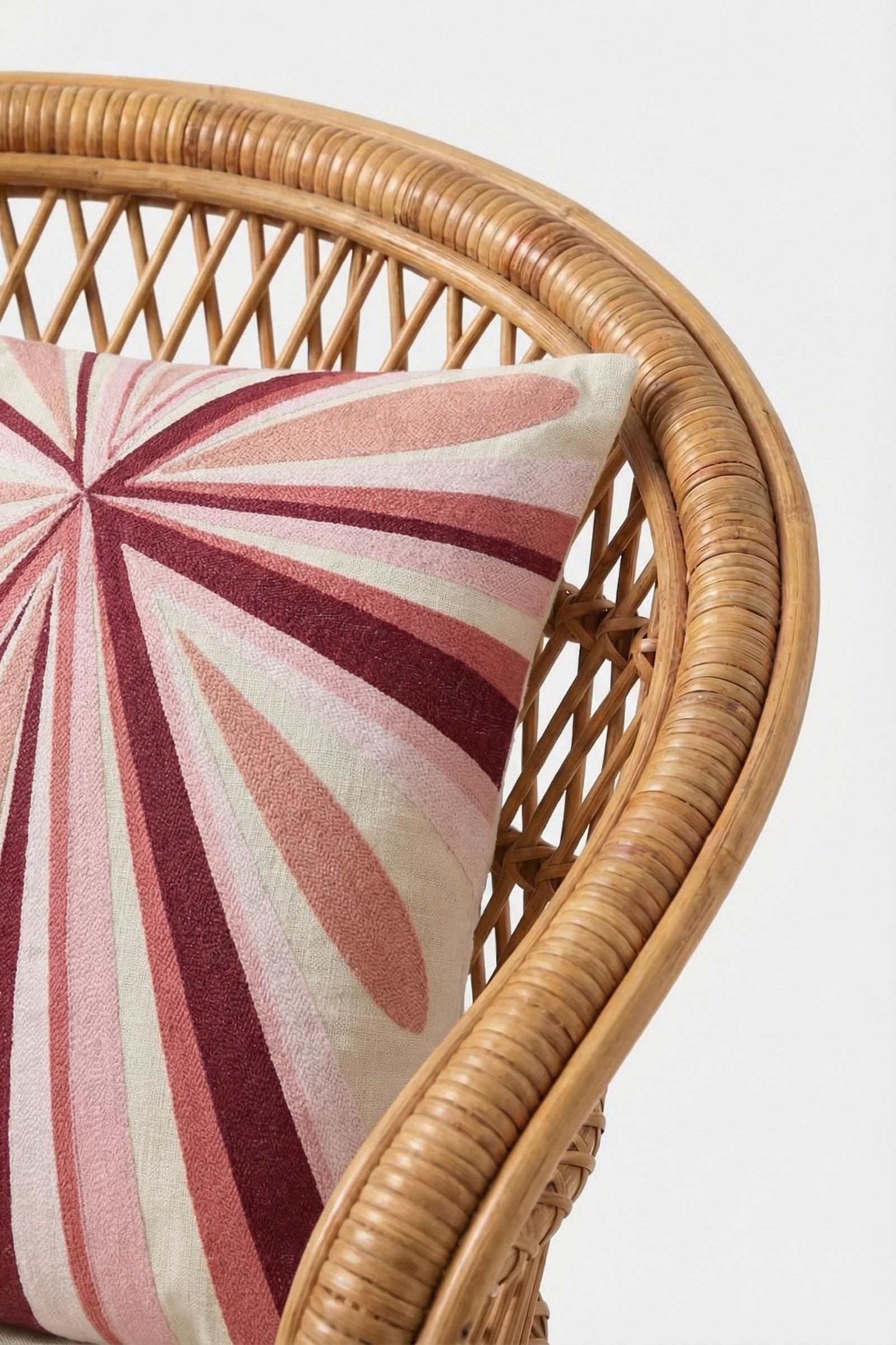 A close-up of a rattan chair with the Shante Embroidered Pillow, featuring a geometric sunburst pattern in pink, red, and cream. The background is plain white.