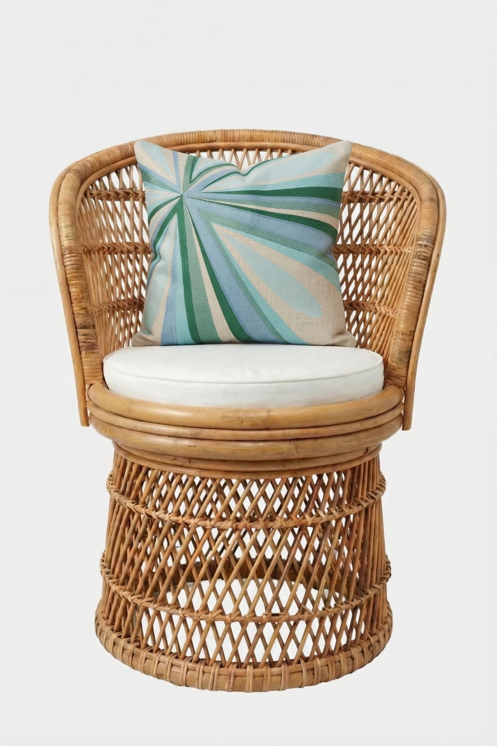A round rattan chair with a crisscross pattern holds a white seat cushion and the Shante Embroidered Pillow, featuring green and blue rays on beige, all set against a plain white backdrop.