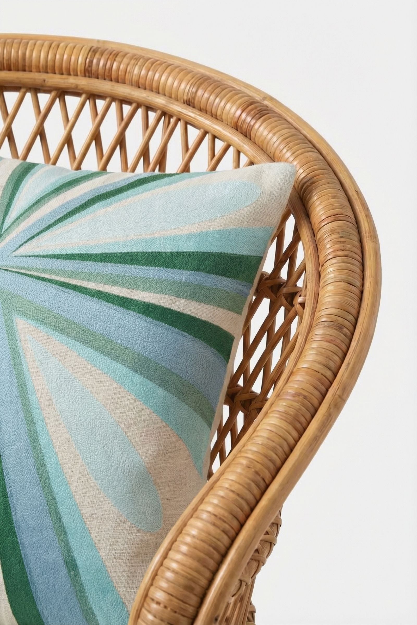 A close-up of a rattan chair with the Shante Embroidered Pillow, showcasing its modern abstract design in green, blue, and cream against a plain white background.
