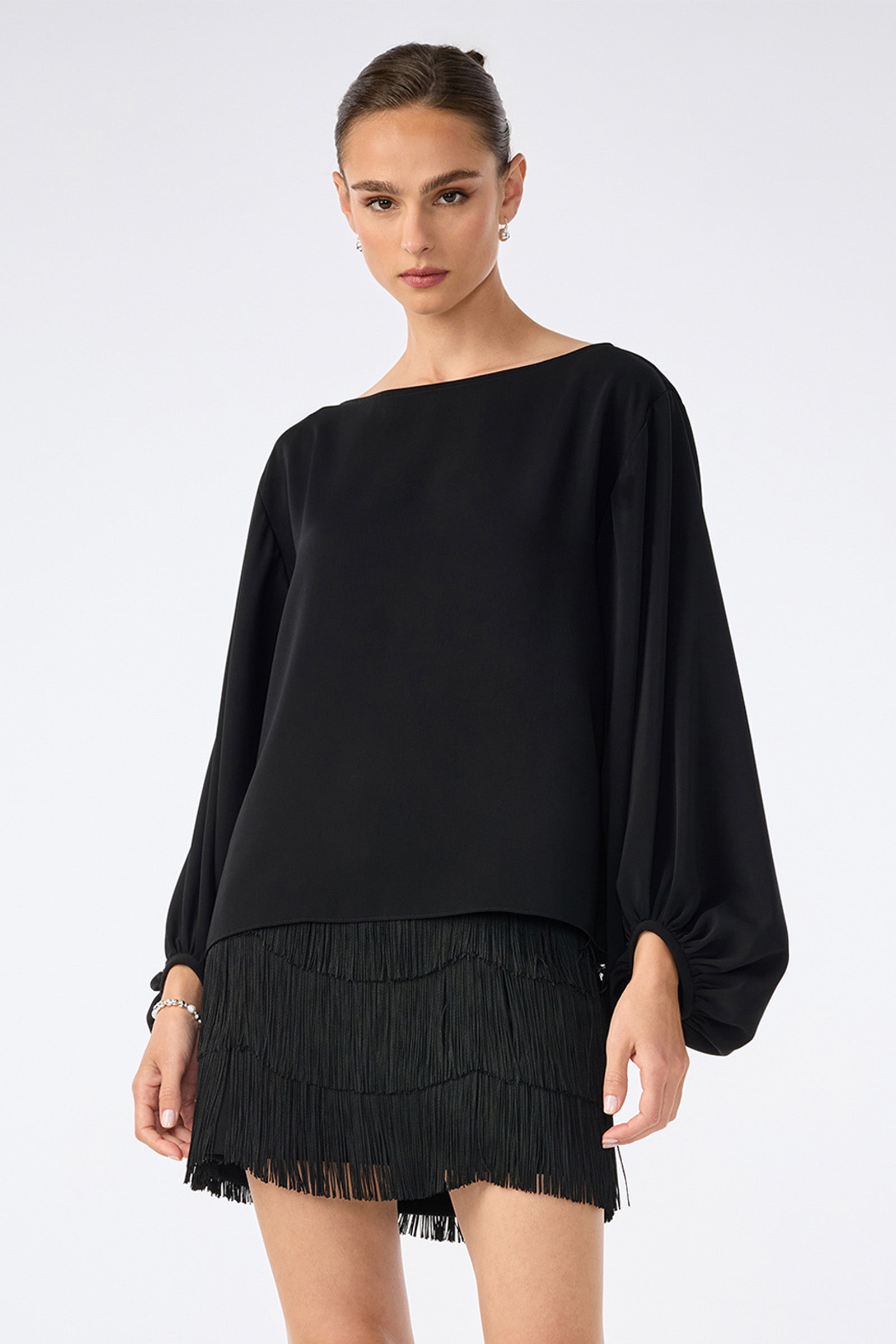 A woman wears the URSA 2 TOP, a black long-sleeved blouse with voluminous sleeves, paired with a short black fringed skirt made from 100% polyester, standing against a plain light background.