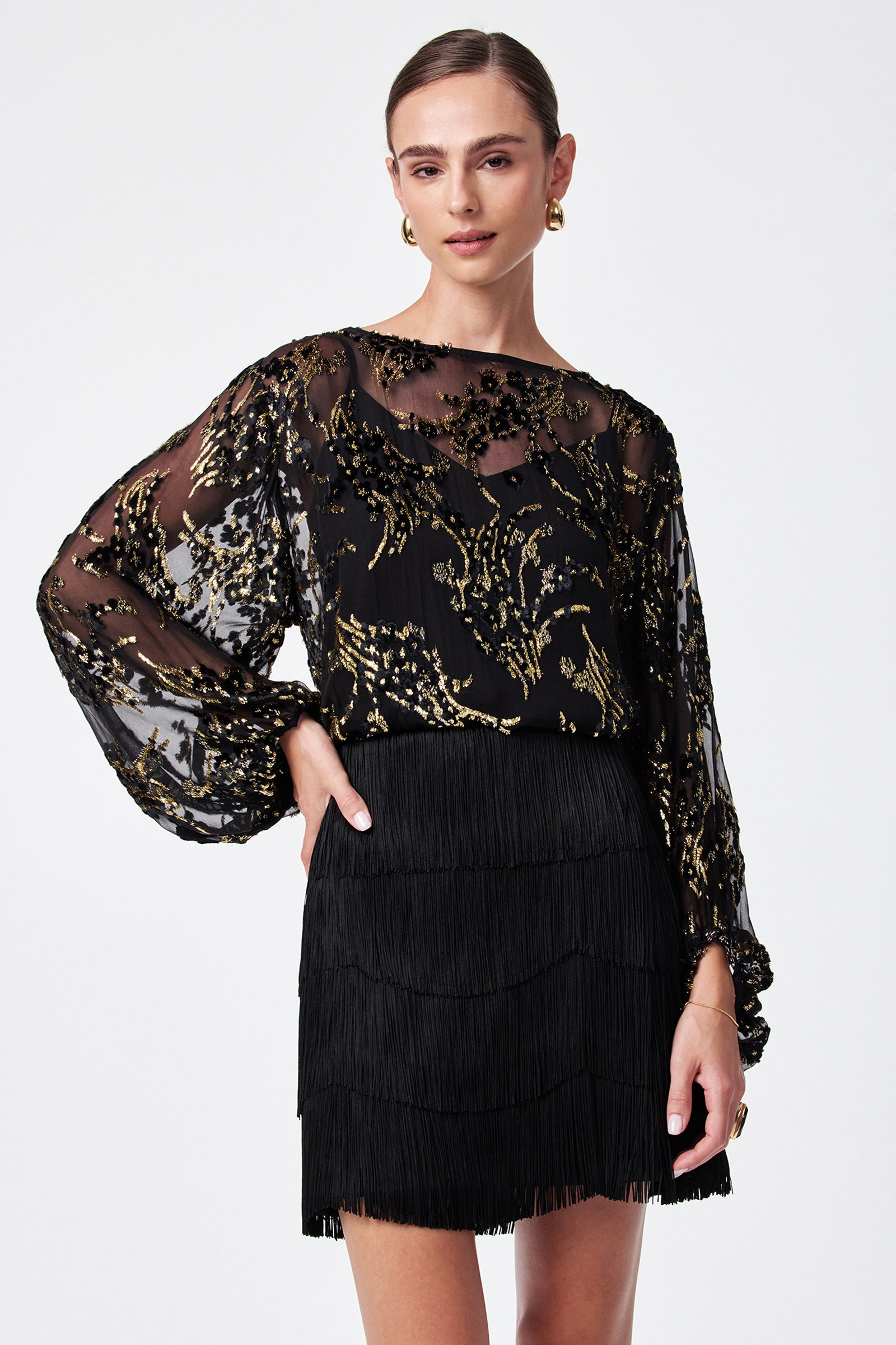 A woman wears the Ursa Metallic Burnout Balloon Sleeve Top, a sheer black blouse with gold floral embroidery and voluminous sleeves, paired with a short black fringed skirt. Her hair is pulled back; she accessorizes with gold earrings and a ring.