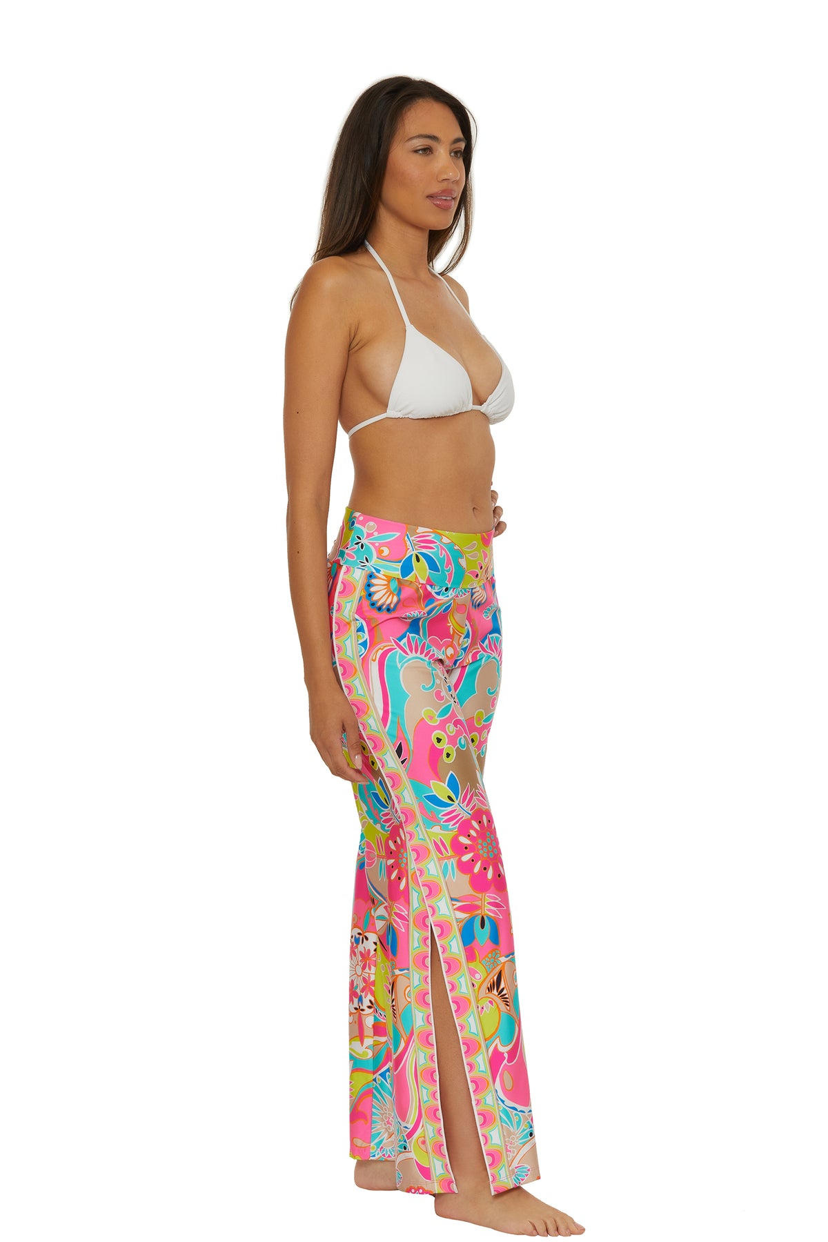 A woman stands barefoot in profile against a plain white background, wearing the Venus Swim Cover-Up Pant—bright, multicolored with a floral pattern and side slits—paired with a white bikini top.