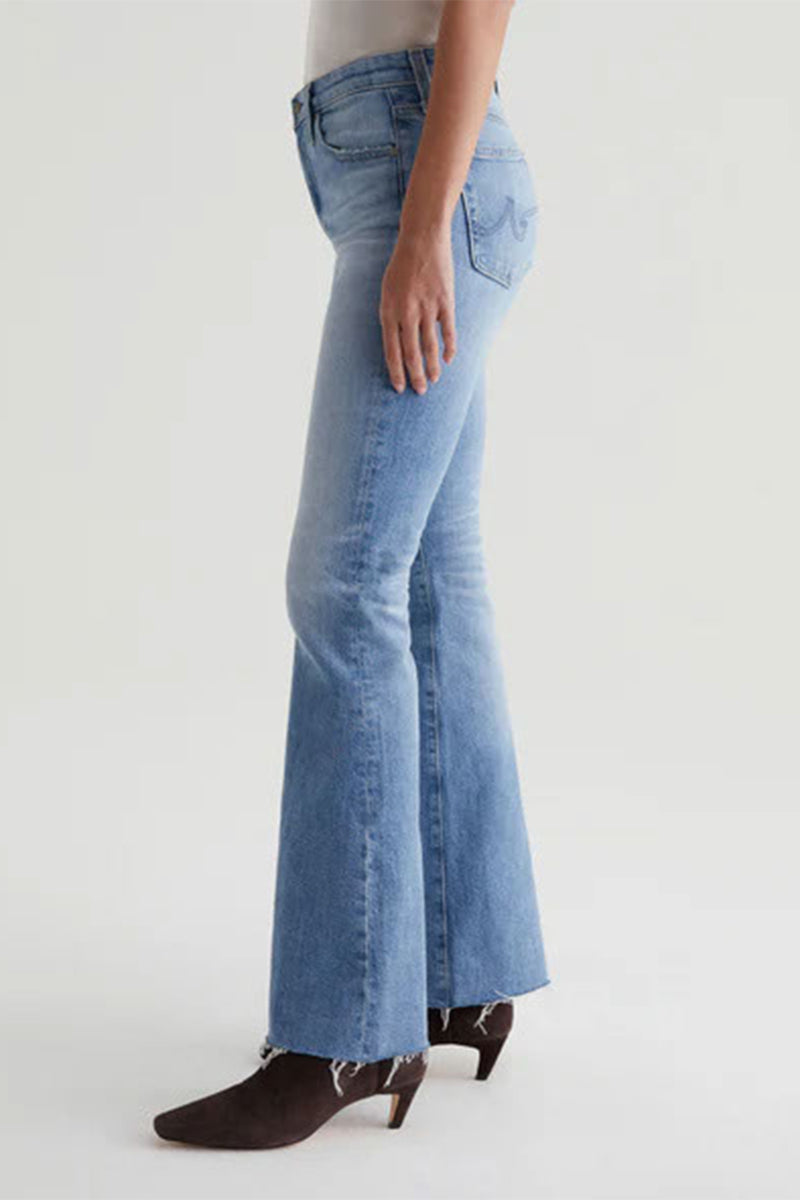 A person wearing AG Womens Farrah Bootcut Blue Jeans—a light wash, high-rise bootcut style with a raw hem—and brown heeled ankle boots, shown from the side against a plain light background.