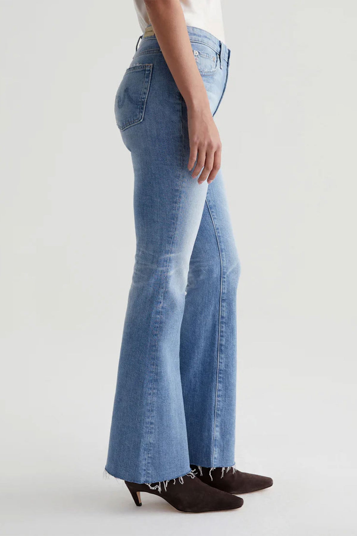 A person stands sideways wearing AG Womens Farrah Bootcut Blue Jean in a light wash with raw hems and brown heeled ankle boots. The background is plain and light-colored.