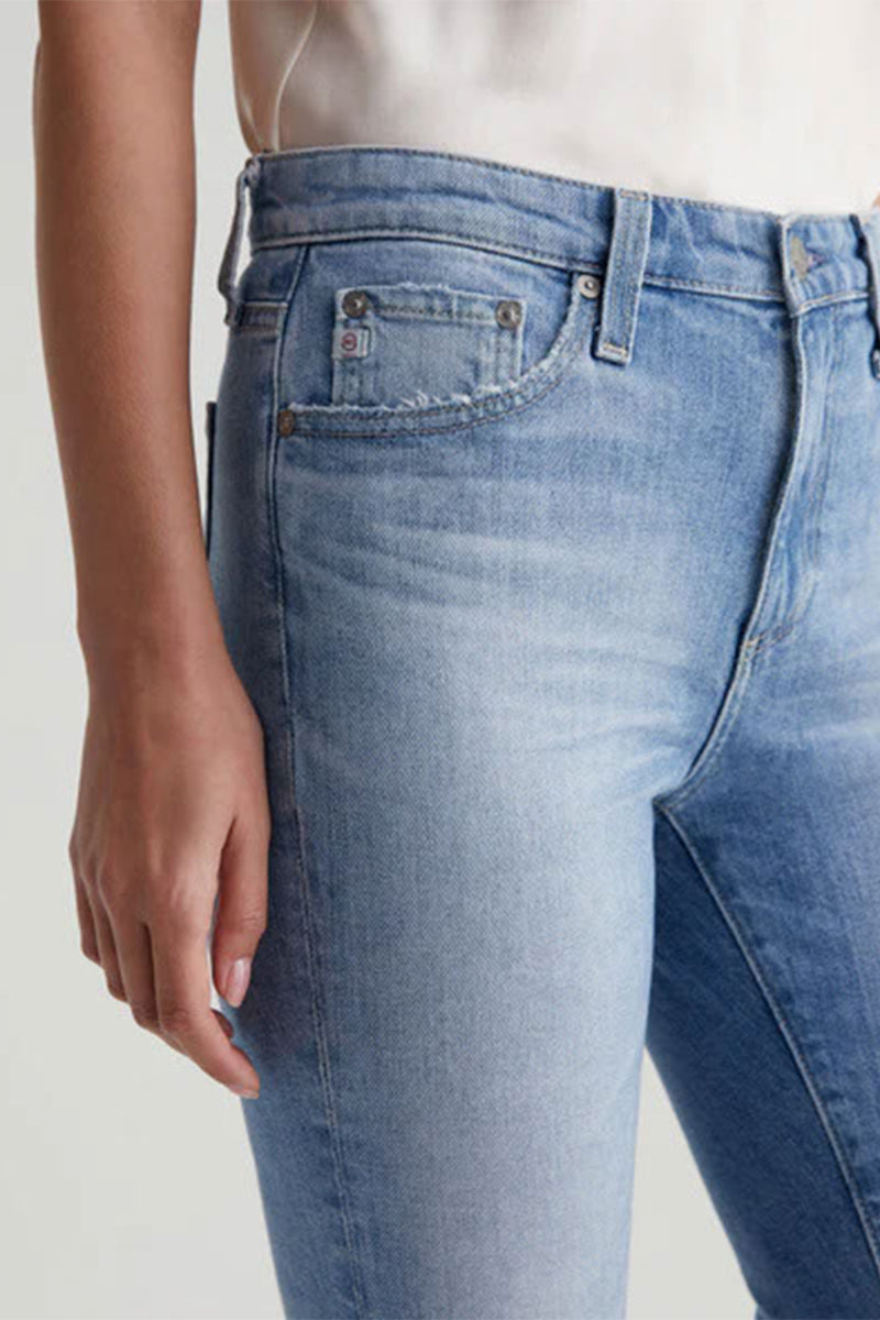 A person wearing AG Womens Farrah Bootcut Blue Jean in light wash, high-rise style, paired with a white top, shown from waist to mid-thigh with one arm resting by their side.