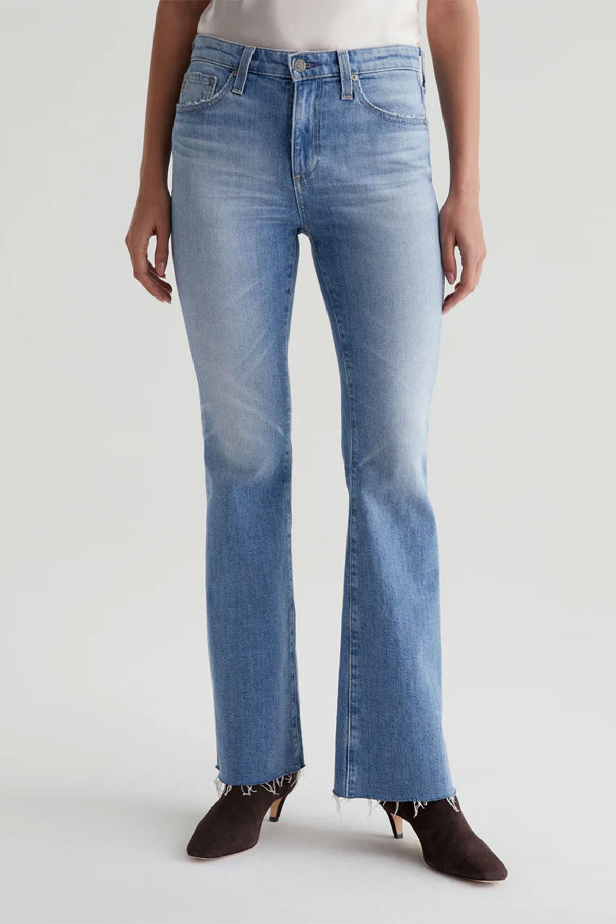 A person in AG Womens Farrah Bootcut Blue Jeans, blue color, with a raw hem and dark heeled ankle boots, stands against a plain light background.