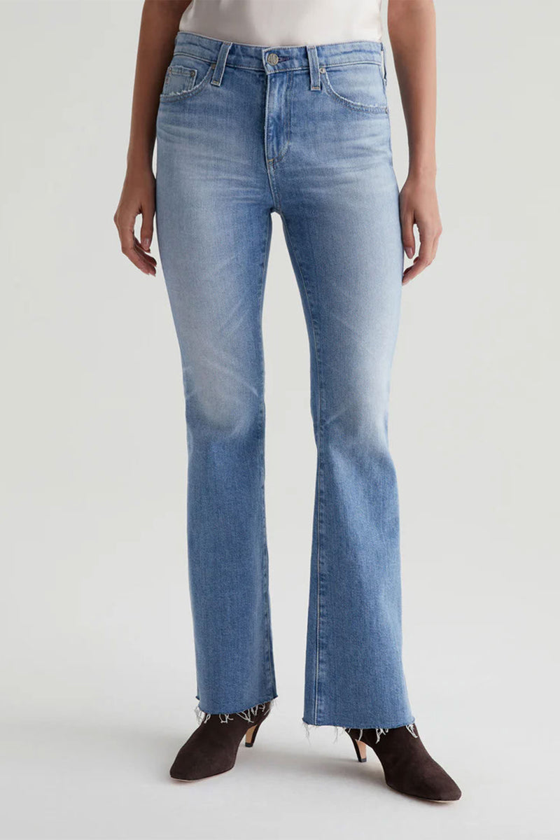 A person in AG Womens Farrah Bootcut Blue Jeans, blue color, with a raw hem and dark heeled ankle boots, stands against a plain light background.
