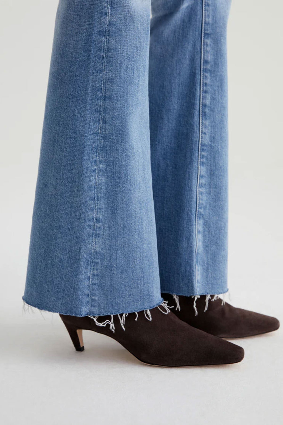 A person wears AG Womens Farrah Bootcut Blue Jeans with a light wash and frayed hem, paired with pointed-toe brown suede kitten heels, standing on a light background.