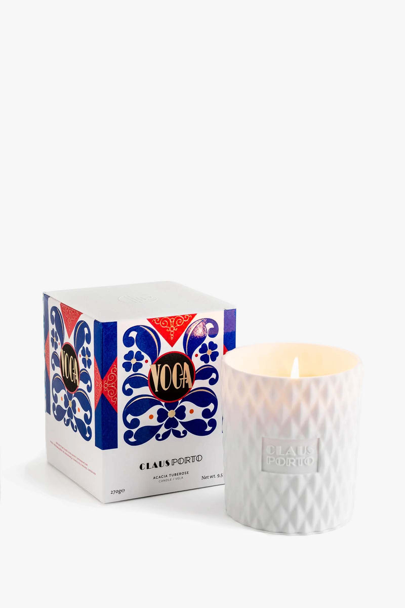 A lit Claus Porto Voga Candle in a textured white glass holder sits next to its decorative box adorned with blue and red patterns.