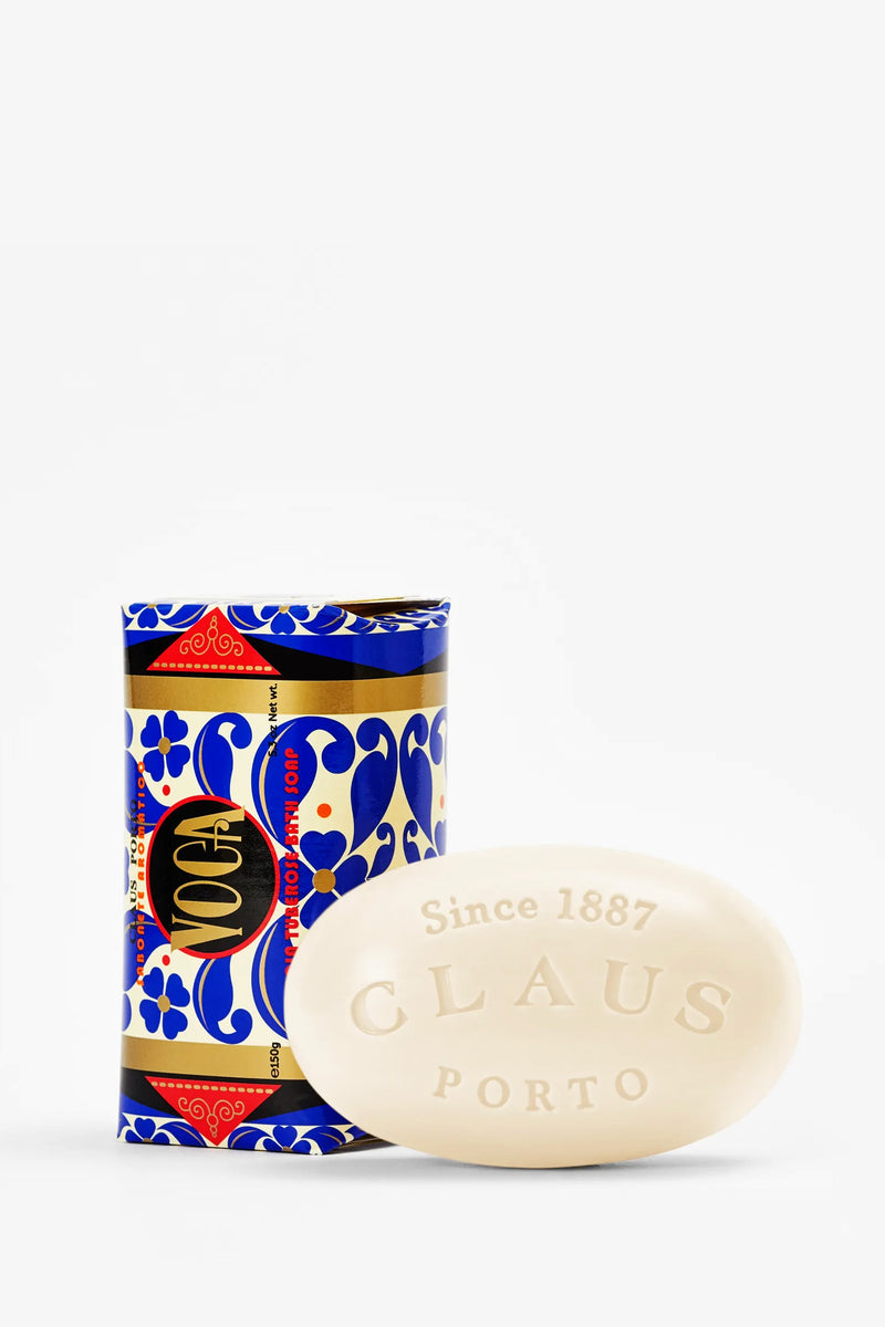 An oval white bar of Claus Porto Voga Soap, embossed with “Since 1887 CLAUS PORTO,” sits in front of a cylindrical decorative box adorned in blue, red, and gold and labeled “Voga.”.