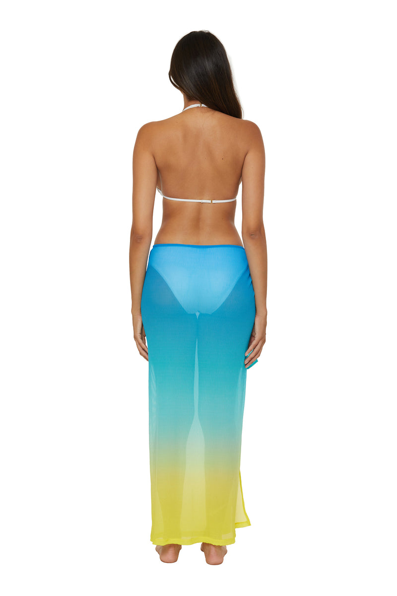 A woman stands barefoot facing away, wearing a white bikini top and the Pareo Swim Cover-Up - West Palm, a sheer maxi skirt that fades from blue at the waist to yellow at the hem. The background is plain white.