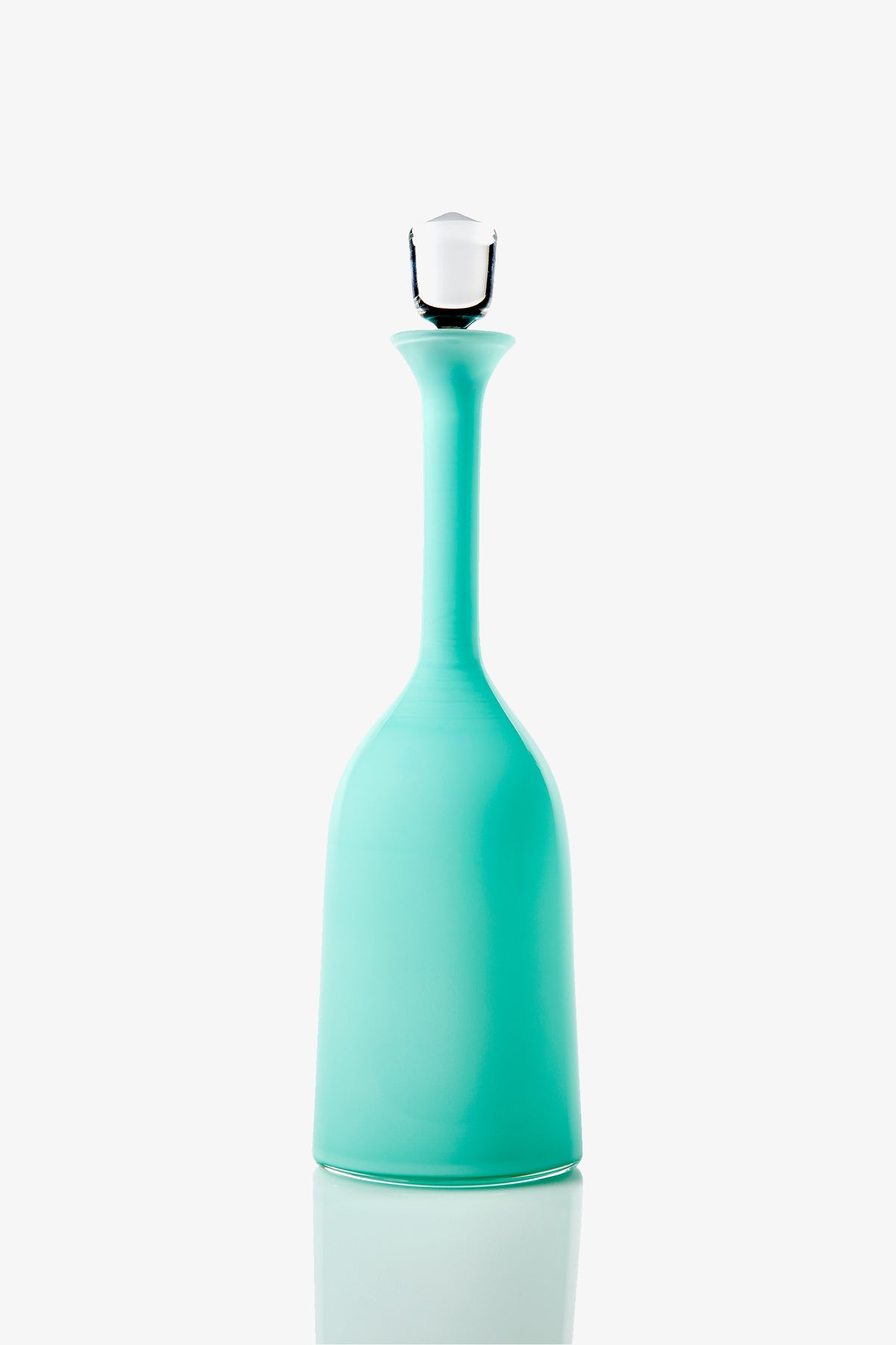 The Wexler Wide Bottle, a tall turquoise glass vessel with a long neck and white stopper, stands upright on a reflective white surface against a plain white background.