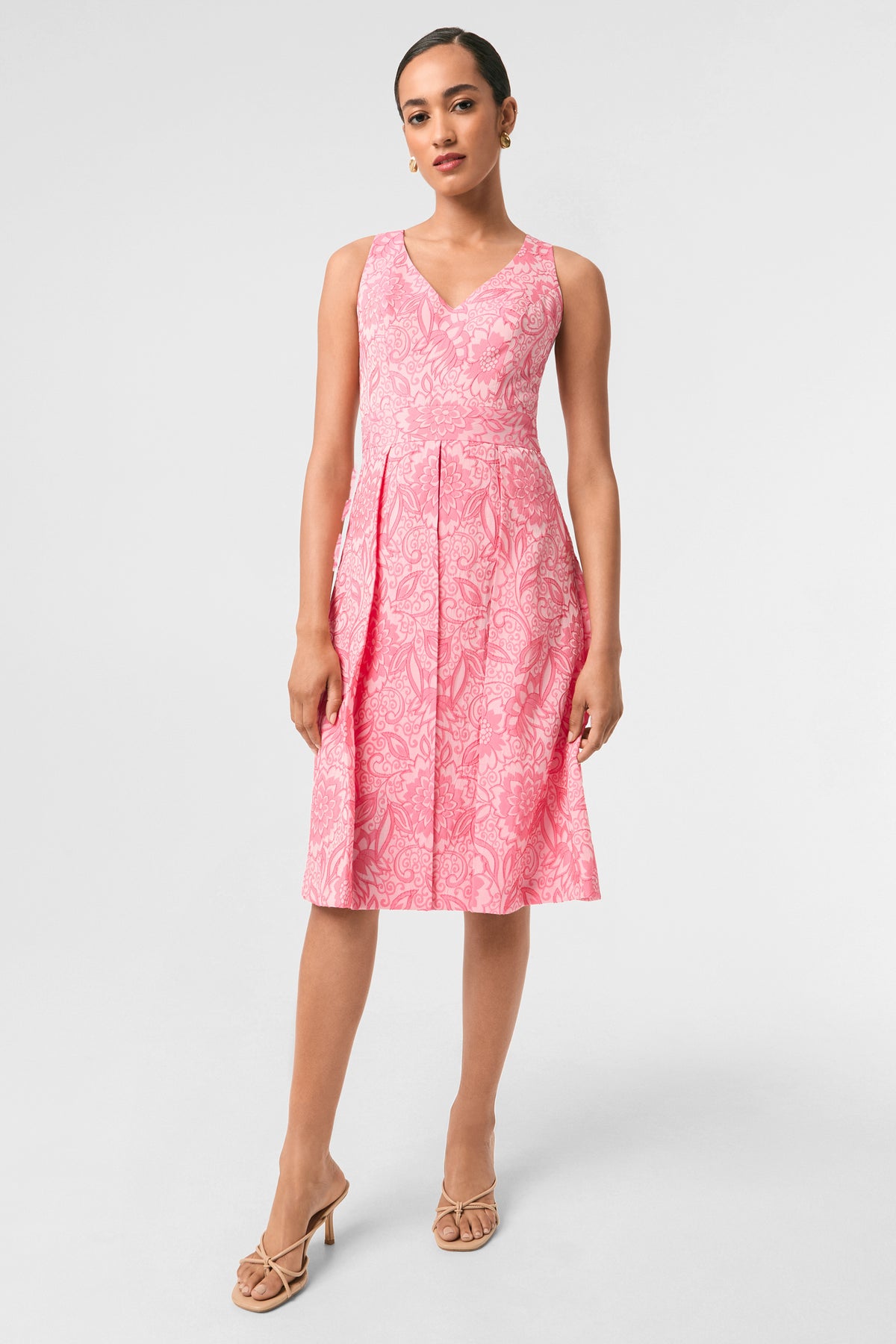 A woman poses against a plain background in the Jayme Sleeveless Dress - Jolie Jacquard in Eden Roc Rose, featuring a V-neckline, knee-length pleated skirt, and nude heeled sandals.