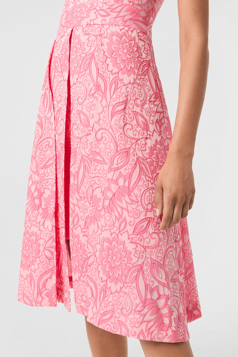 Someone wearing the Jayme Sleeveless Dress - Jolie Jacquard, a pink and white knee-length dress with a floral and leaf pattern, shows their right arm and part of their torso against a plain light background.