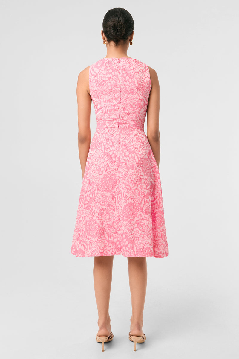 A woman faces away in the Jayme Sleeveless Dress - Jolie Jacquard, a knee-length pink dress with a white floral pattern. Her hair is tied back, she wears beige heeled sandals, and stands against a plain light gray background.