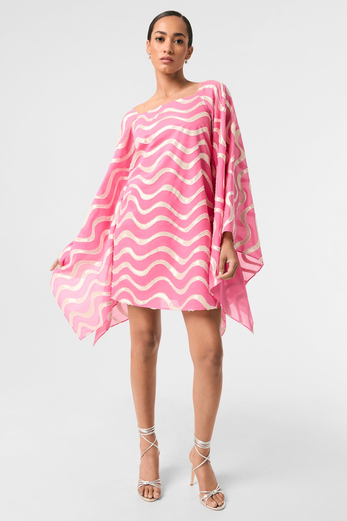 Woman in the Aqualane Side Flounce Dress - Pinku, a loose mini dress with pink and white wavy patterns and wide sleeves, stands with one leg forward and arms extended, styled with strappy silver heels against a plain background.