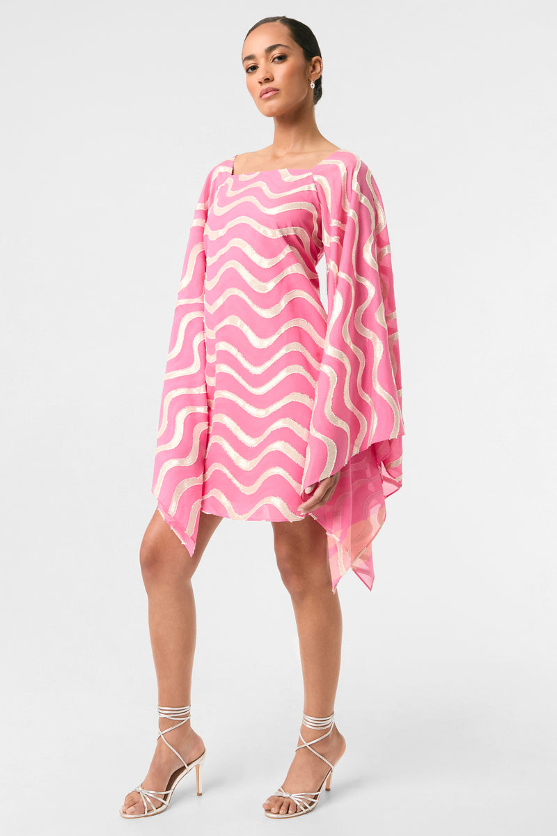 A woman poses confidently in the Aqualane Side Flounce Dress - Pinku, a pink and white wavy patterned mini dress with wide, flowing sleeves, styled with strappy high-heeled sandals against a white background.