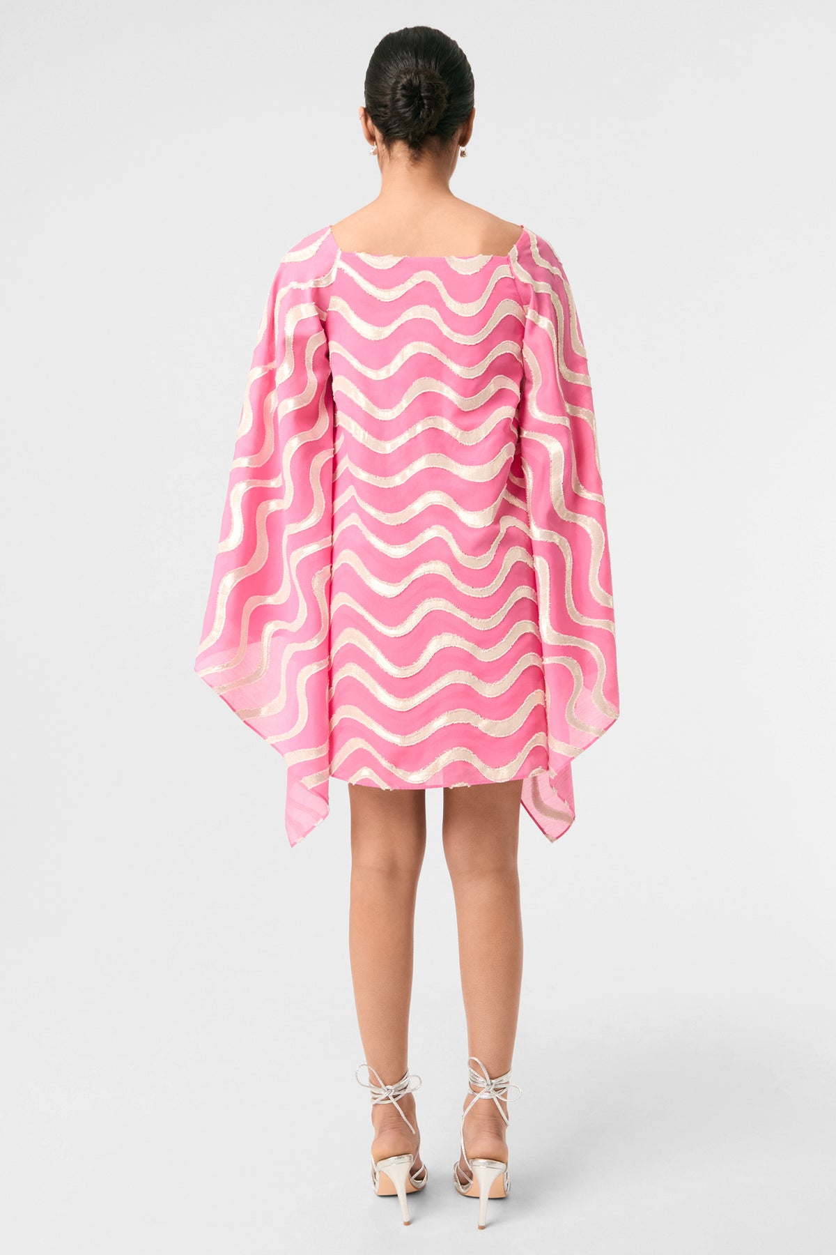 A woman faces away, wearing the Aqualane Side Flounce Dress - Pinku with pink and white wavy stripes, long flowy sleeves, open sides, her hair tied back, and white high-heeled sandals against a plain background.