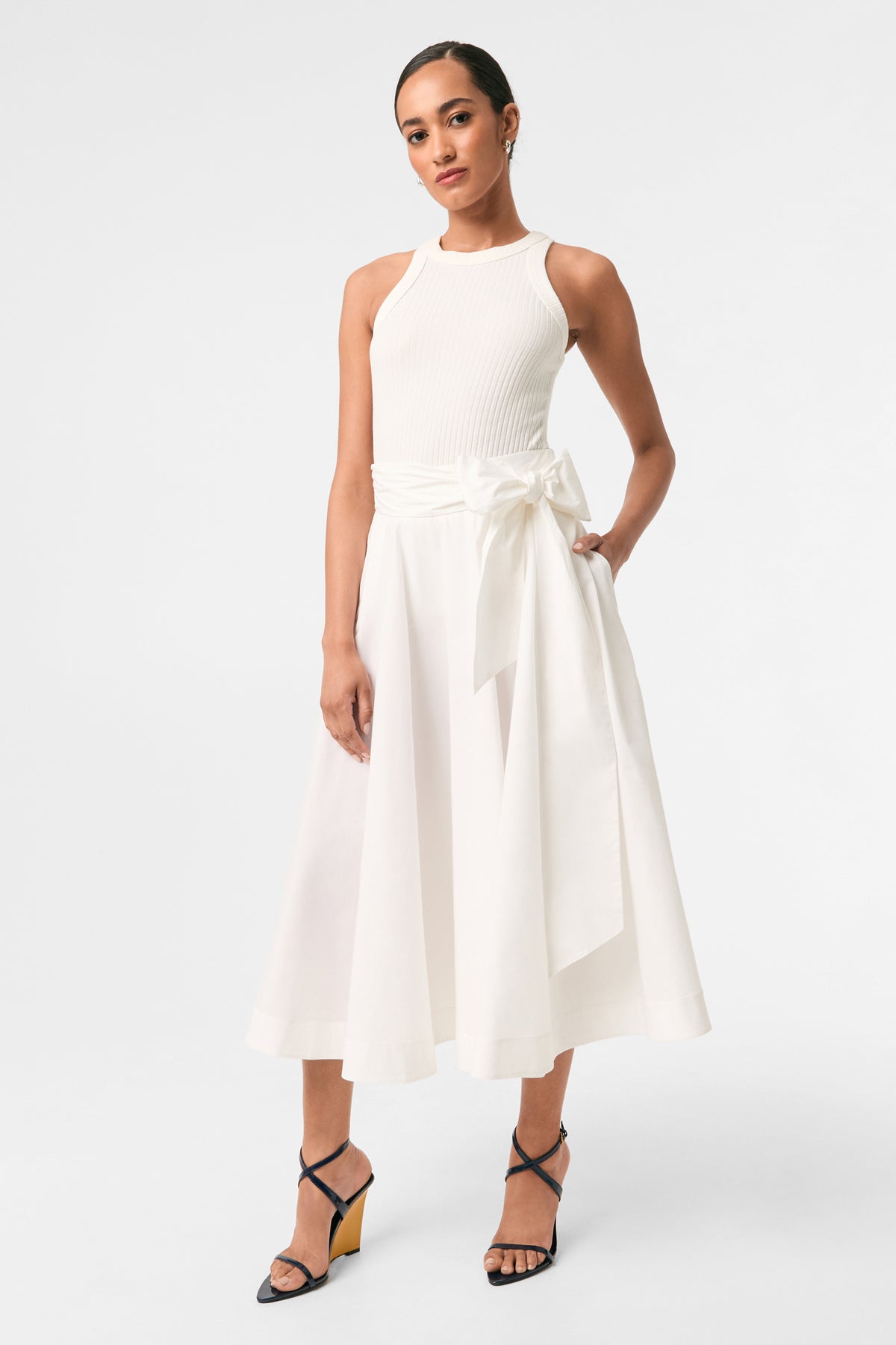 A woman stands against a plain background wearing a sleeveless, fitted white top and the Amano Bow-Tie Midi Skirt, paired with black strappy wedge heels.