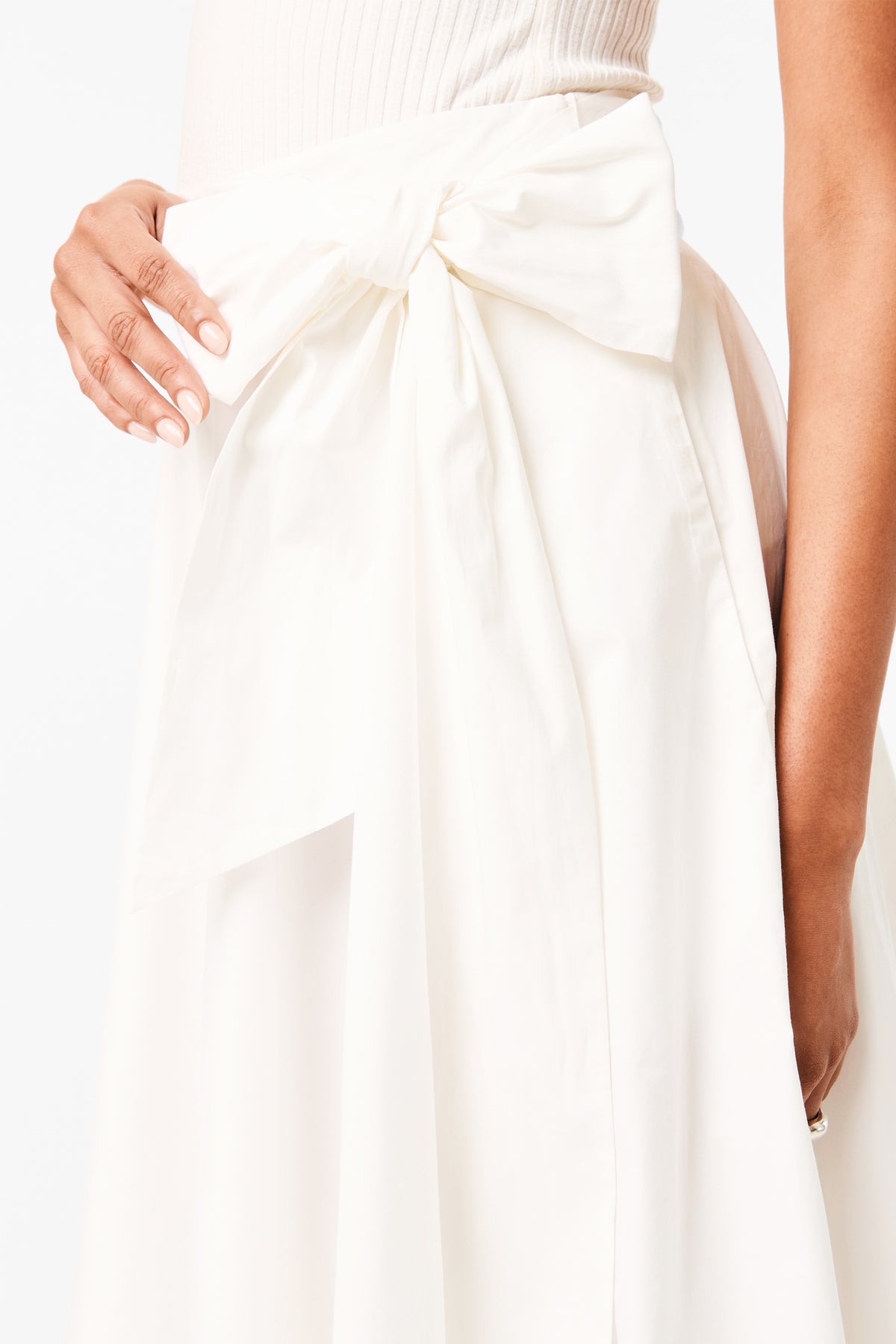 A woman‚Äôs hand rests on her waist, highlighting the large bow of the Amano Bow-Tie Midi Skirt. She pairs it with a white, sleeveless top against a plain white background.