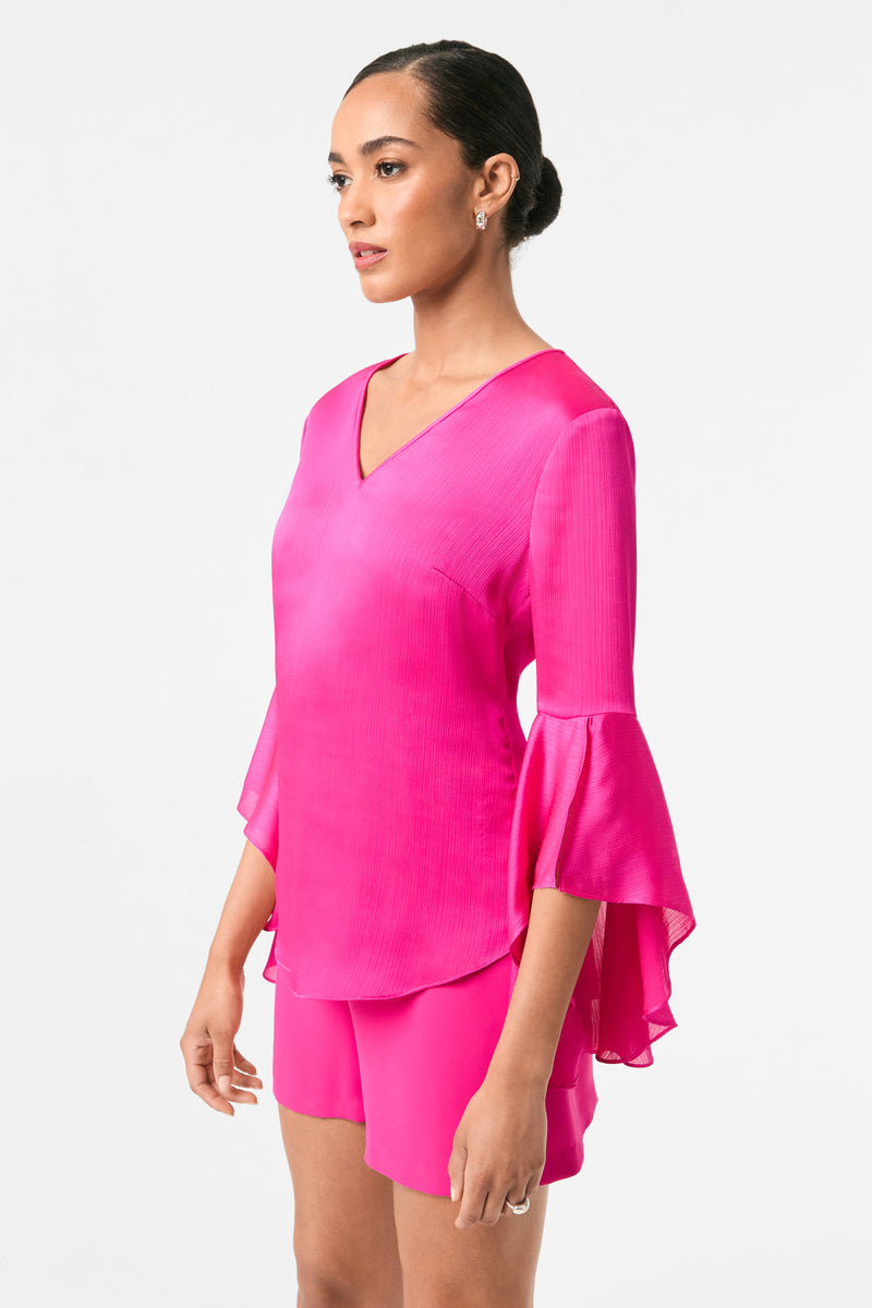 A woman stands against a plain white background, wearing the Yuki Flounce-Sleeve Top in bright pink with flared sleeves and matching shorts. Her hair is styled in a neat bun.