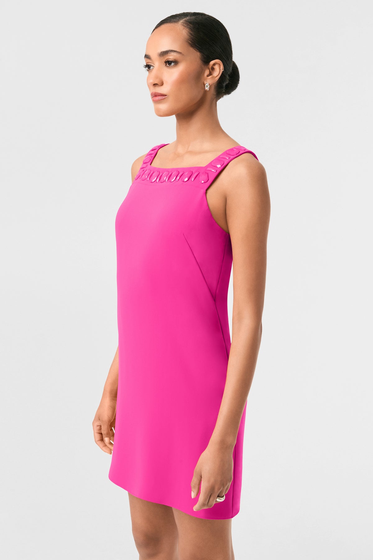 A woman stands in profile against a plain background, wearing the Yokuna Jewel Neck Mini Dress in bright pink, featuring a sleeveless design, square neckline with decorative collar details. Her hair is styled in a sleek bun and she has a calm expression.