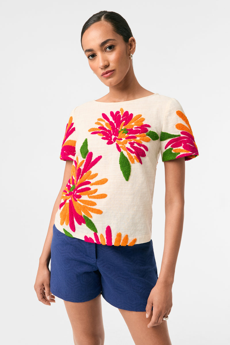 A person wears the Siona Embroidered Flowers Top in Riku Embroidery, featuring bold pink, orange, and green floral designs, paired with dark blue shorts against a plain white background.