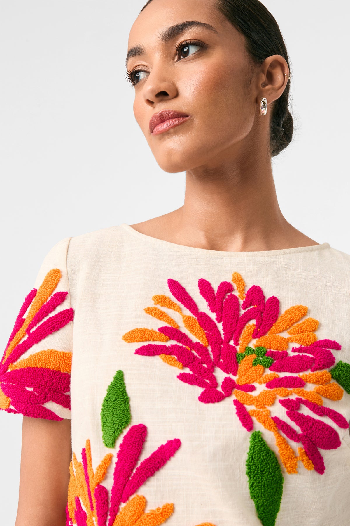 A woman with slicked-back hair models the Siona Embroidered Flowers Top, a cream-colored piece adorned with bold, textured pink, orange, and green floral embroidery. She looks confidently to the side against a plain light background.