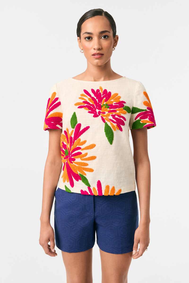 A woman faces forward wearing the Siona Embroidered Flowers Top, featuring cream fabric with bright pink, orange, and green floral designs, paired with navy blue shorts. Her dark hair is pulled back as she poses against a plain light background.