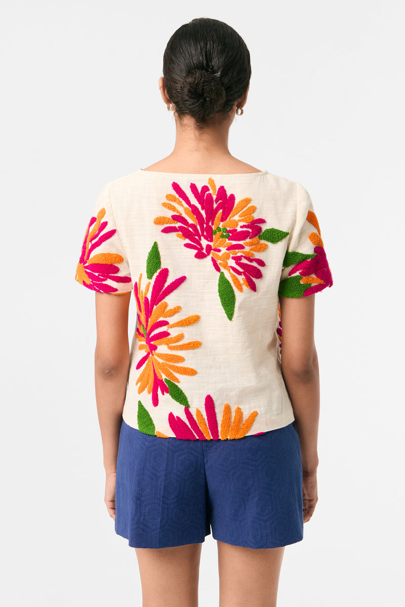 A woman with her hair in a low bun faces away from the camera, wearing the Siona Embroidered Flowers Top‚Äîa cream blouse with vibrant pink, orange, and green floral embroidery‚Äîpaired with blue shorts.