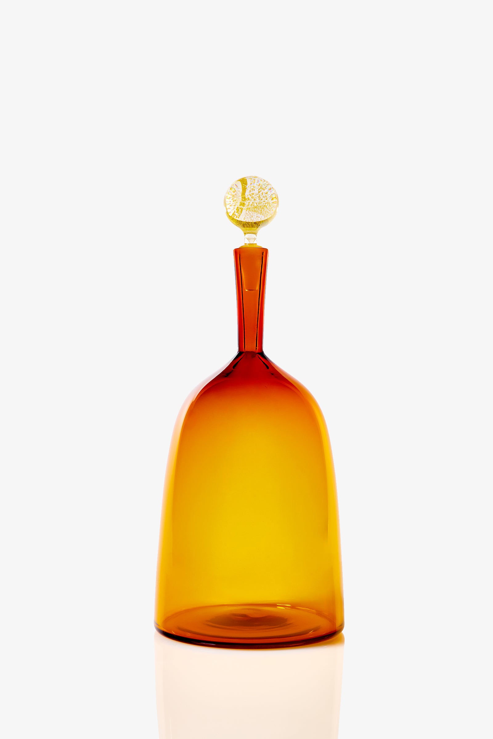 The Wide Bottle Carmella Barware is a tall, amber glass bottle with a wide base, narrow neck, and round gold-patterned stopper, shown against a plain white background.