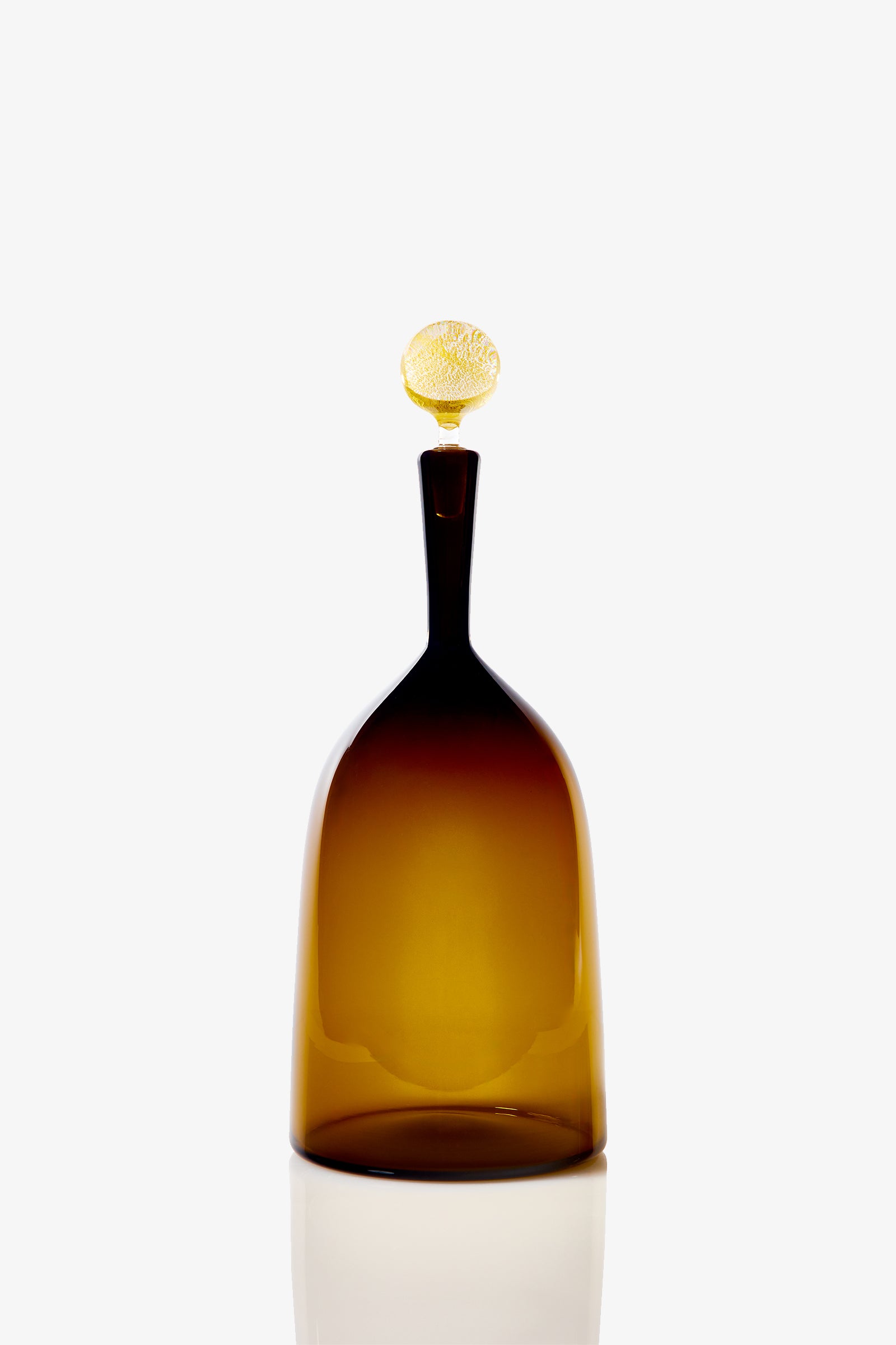 The Wide Bottle Carmella Barware is a tall, translucent amber glass bottle with a narrow neck and a textured, round stopper, displayed against a plain white background.