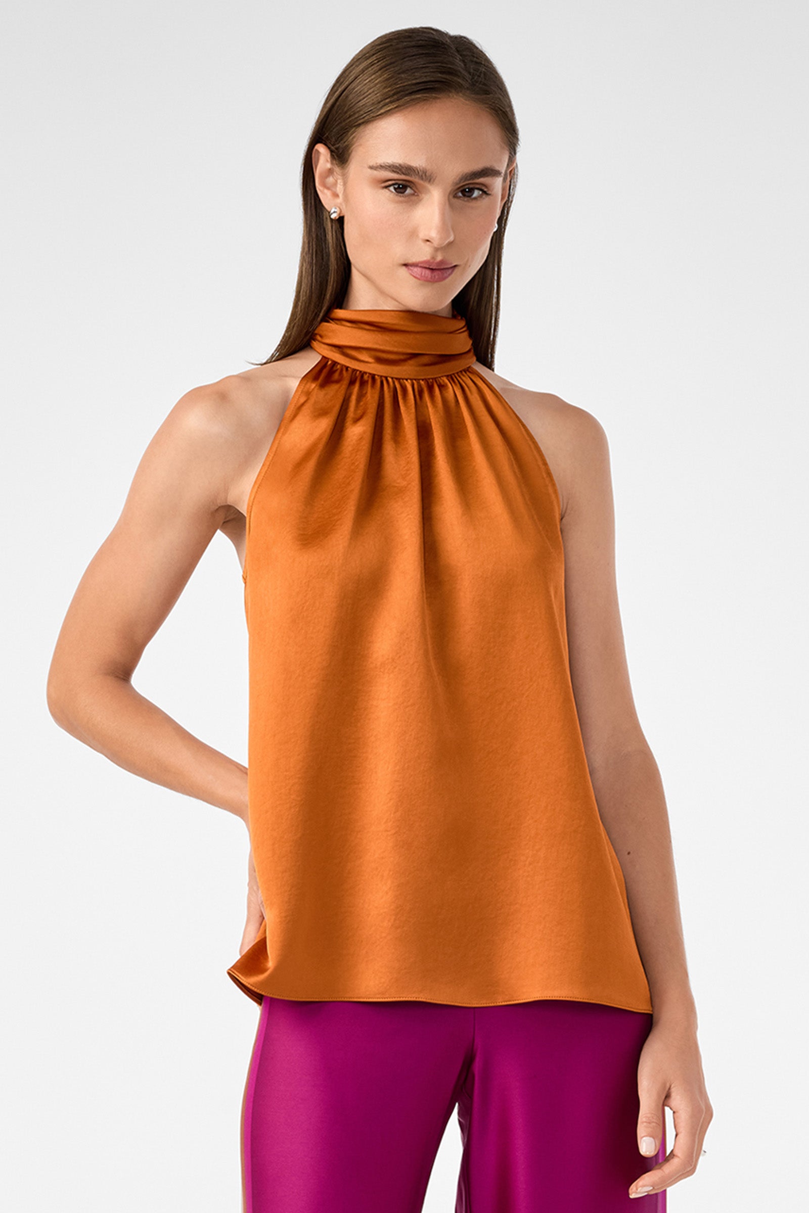 A woman with straight brown hair wears the orange Zana Halter Top and magenta pants, standing against a plain light gray background.