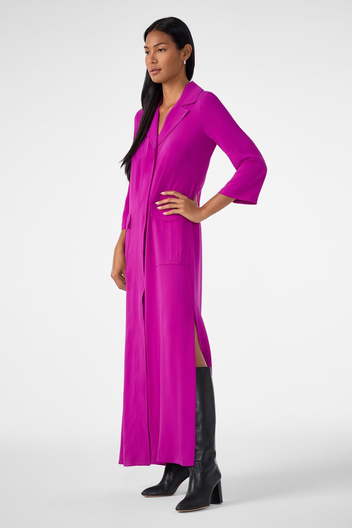A woman models the Annecy Dress—a magenta, long-sleeve, collared button-down with front pockets and side slits—paired with knee-high black boots. She poses with one hand on her hip against a plain white background.