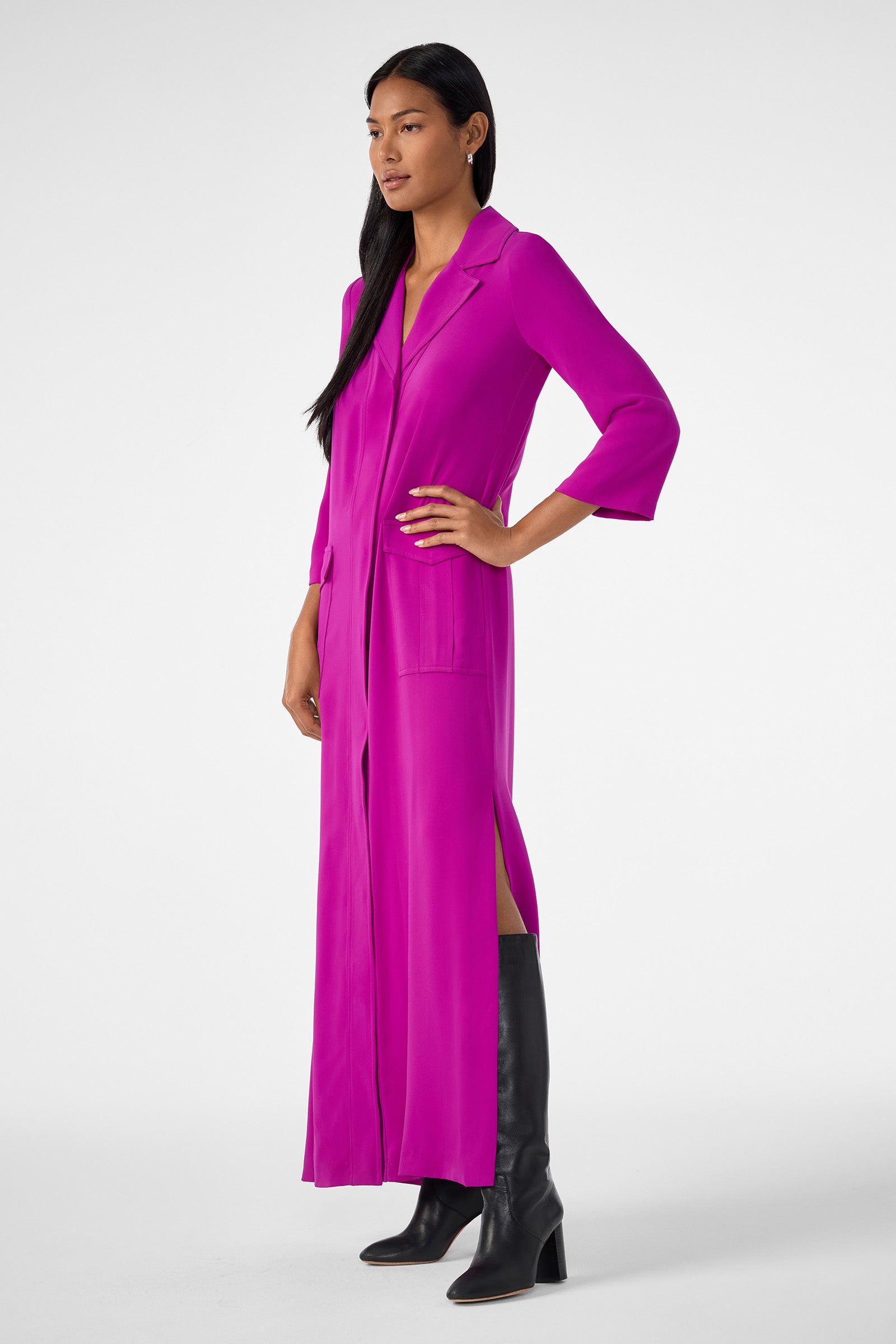 A woman models the Annecy Dress—a magenta, long-sleeve, collared button-down with front pockets and side slits—paired with knee-high black boots. She poses with one hand on her hip against a plain white background.